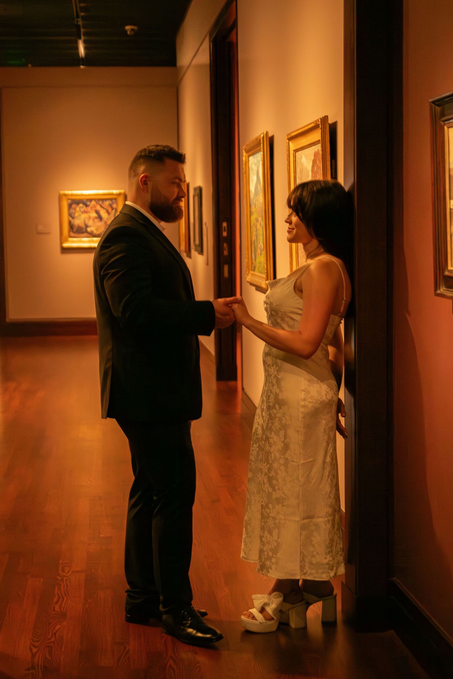 A couple explores an art gallery together, walking through warmly lit corridors with framed artwork.