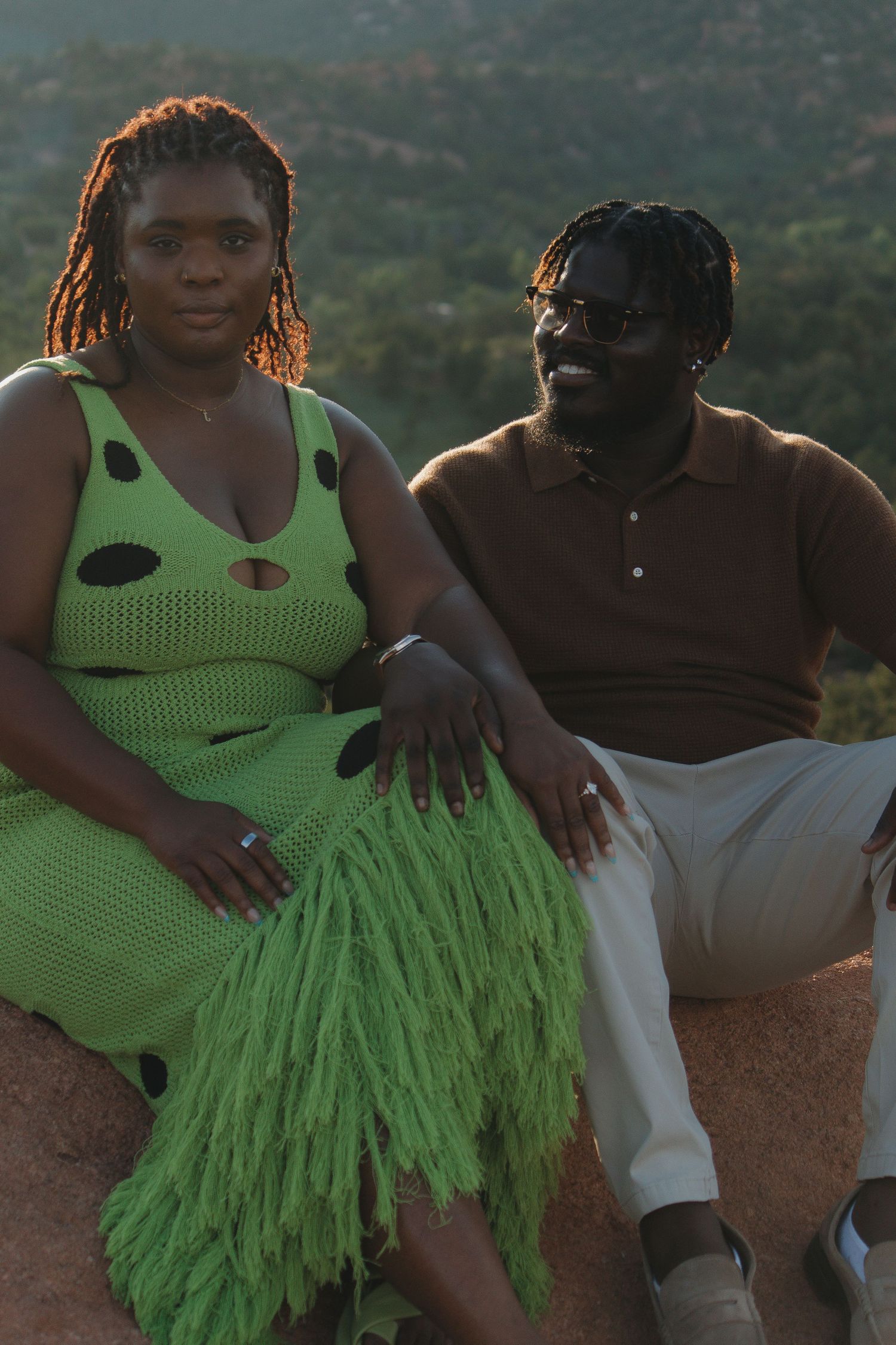 A couple sits together outdoors wearing a lime green polka dot dress and white outfit against a mountainous background.