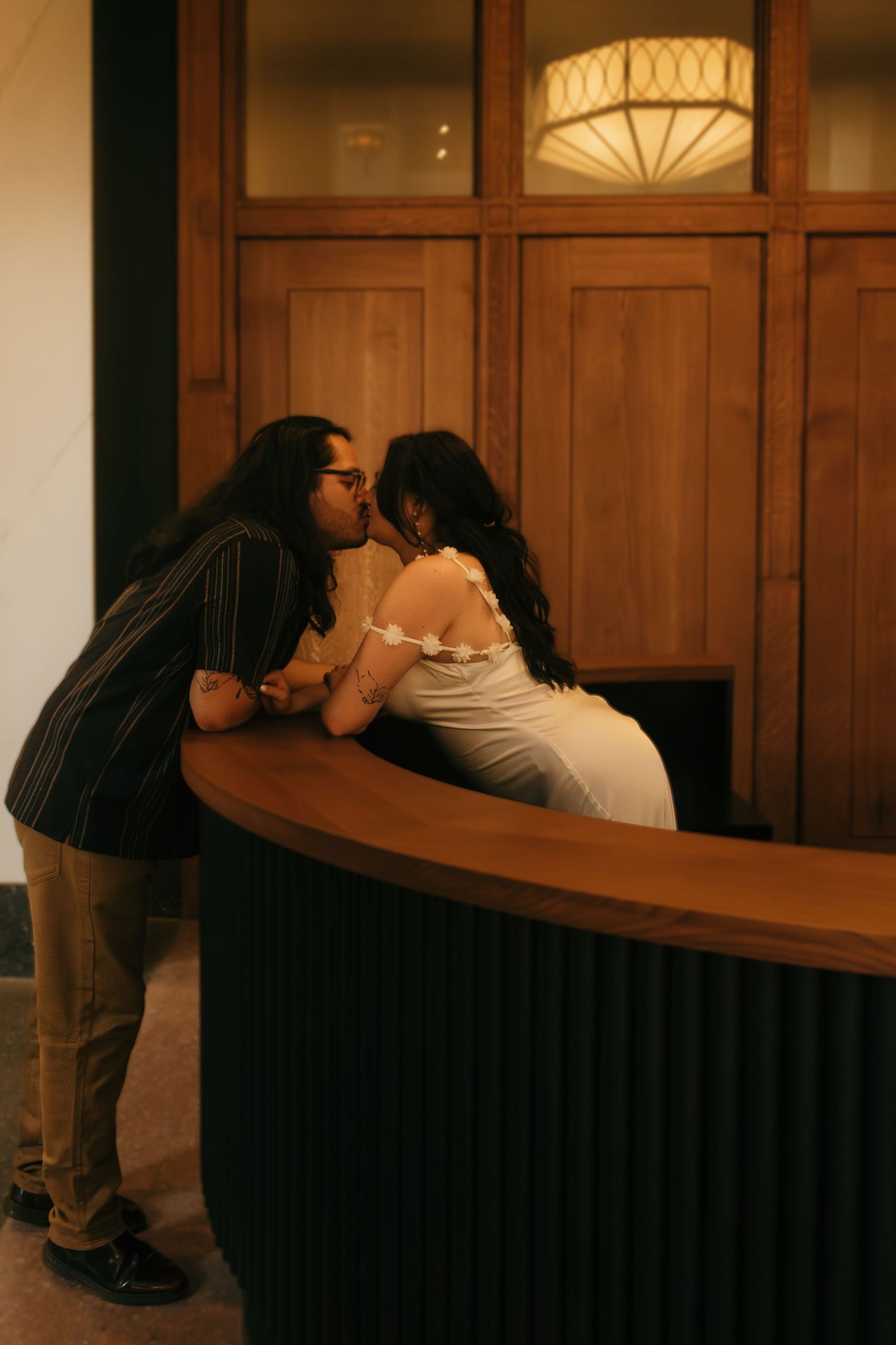 A couple shares a tender moment in front of an elegantly lit curved desk with wood paneling.