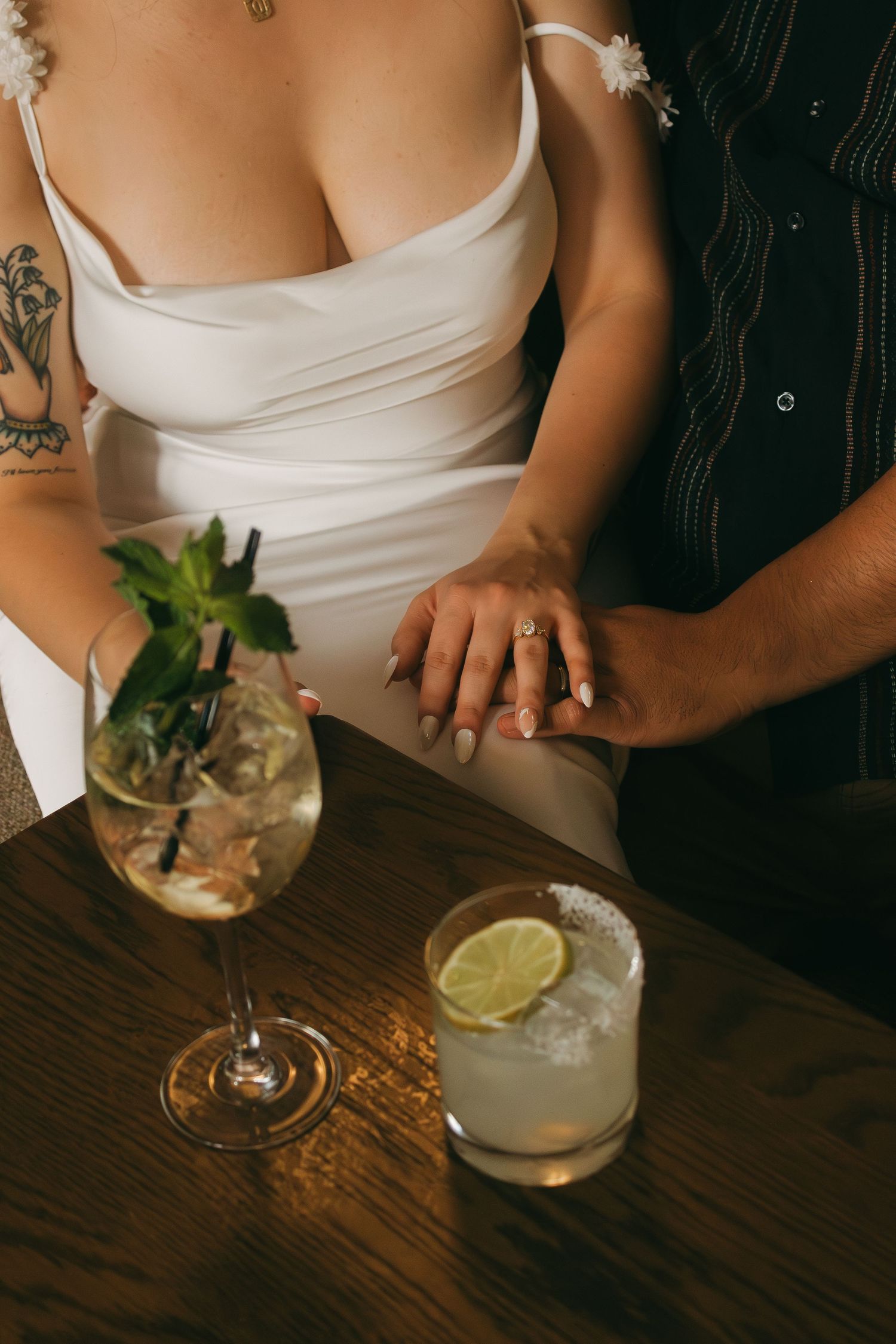 A couple's hands rest together on a wooden table with cocktail glasses and lime garnish.
