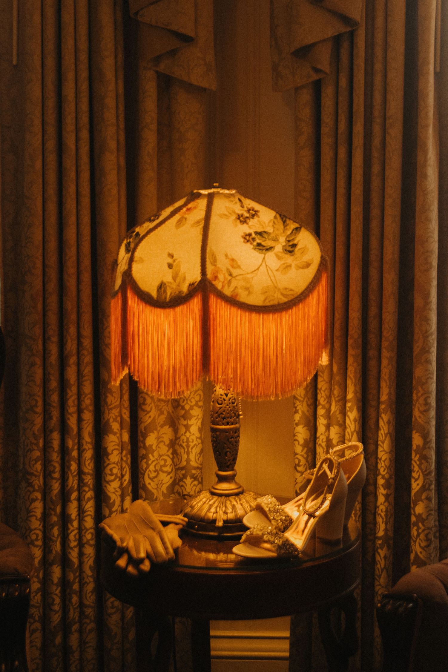 Vintage table lamps with ornate orange fringed shades cast a warm glow through elegant curtains in a dimly lit room.