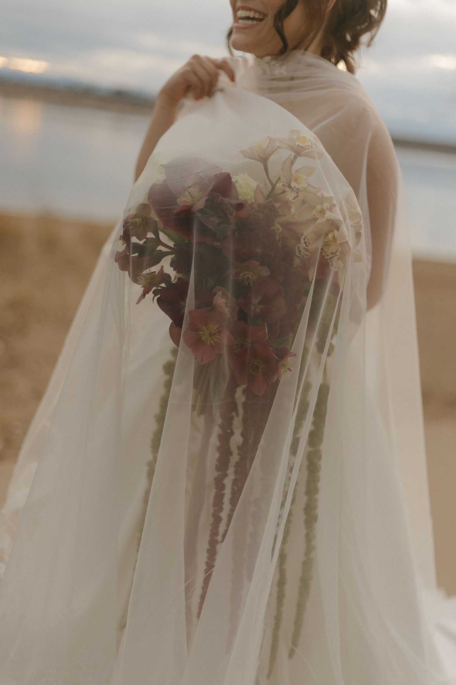A close-up view of a bridal bouquet wrapped in delicate white tulle fabric against a beach backdrop at sunset.