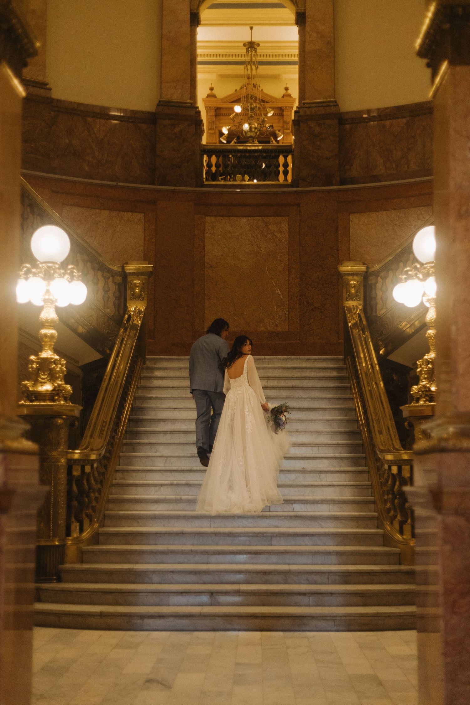 A bride in a white wedding dress descends a grand marble staircase with ornate gold railings in an elegant venue.