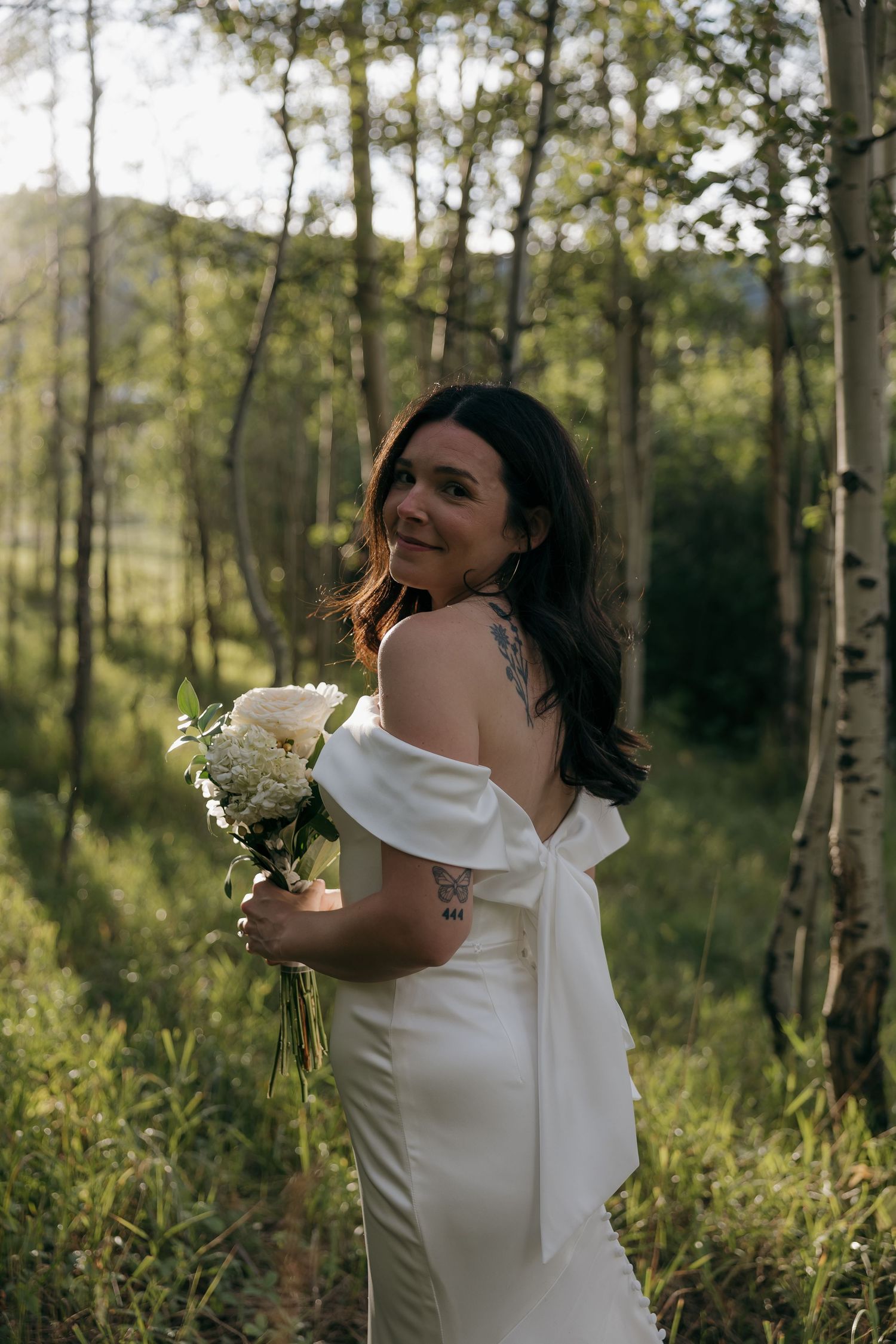 Bride in off-shoulder gown with tattoo holding white bouquet in birch forest.