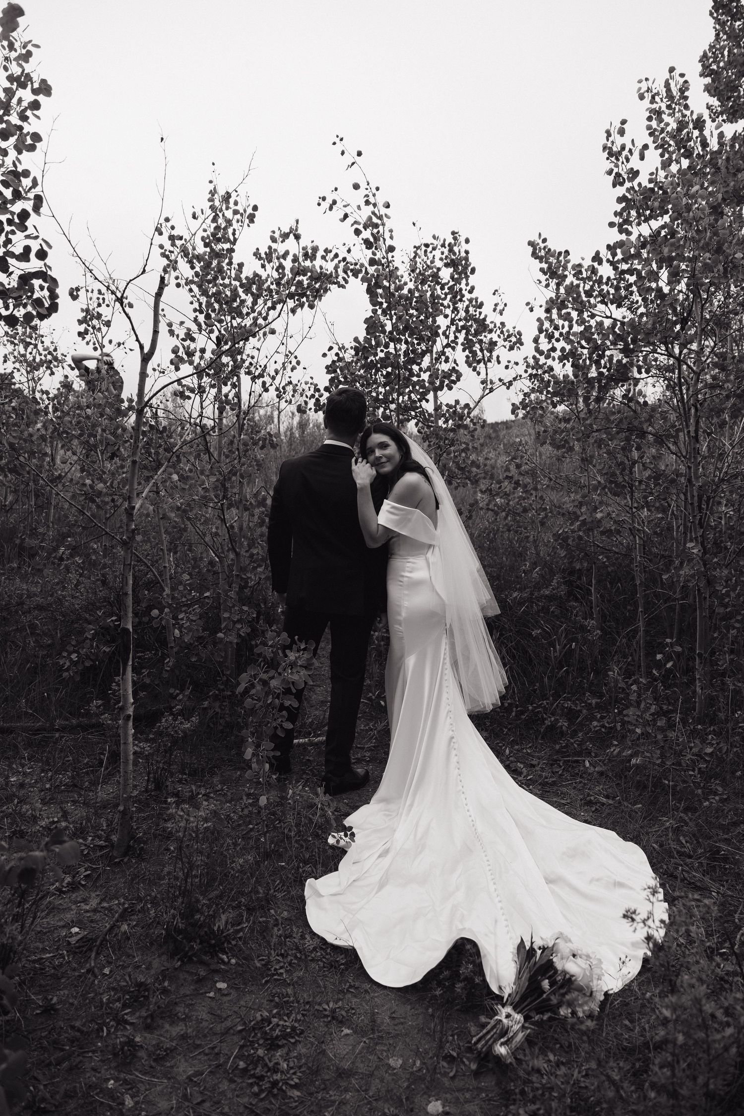 Black and white photo of bride and groom embracing in a forest, brides long train flowing on the ground.