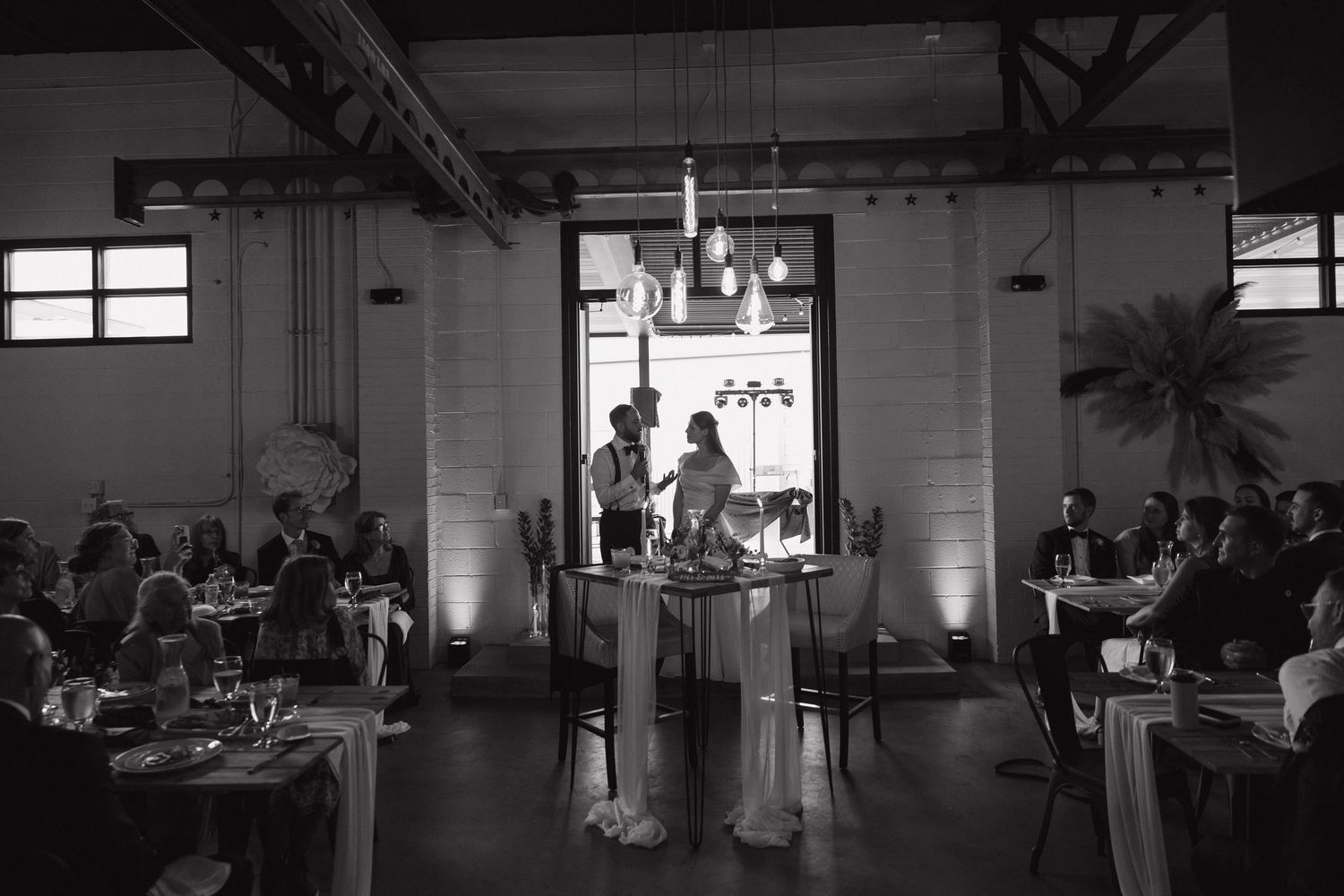 Black and white photo of bride and groom seated at sweetheart table during wedding reception.