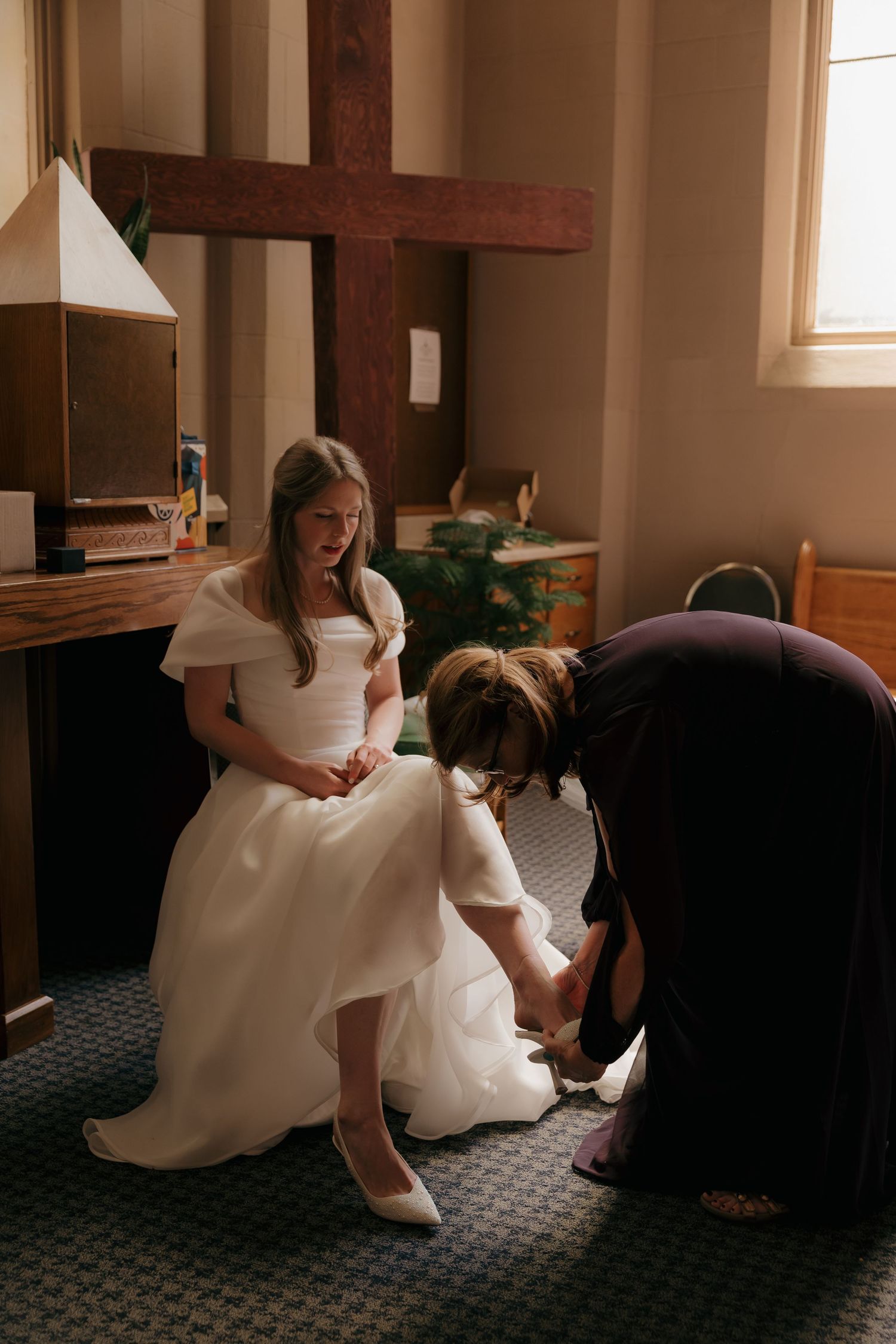Bride in white gown getting shoes put on by helper inside a church before wedding ceremony.