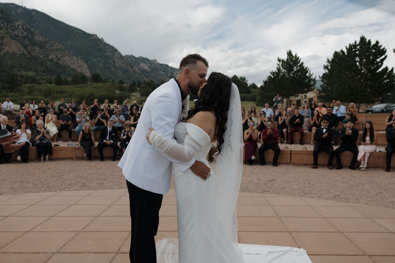 Bride and groom share first kiss at outdoor wedding ceremony with mountain backdrop and seated guests.