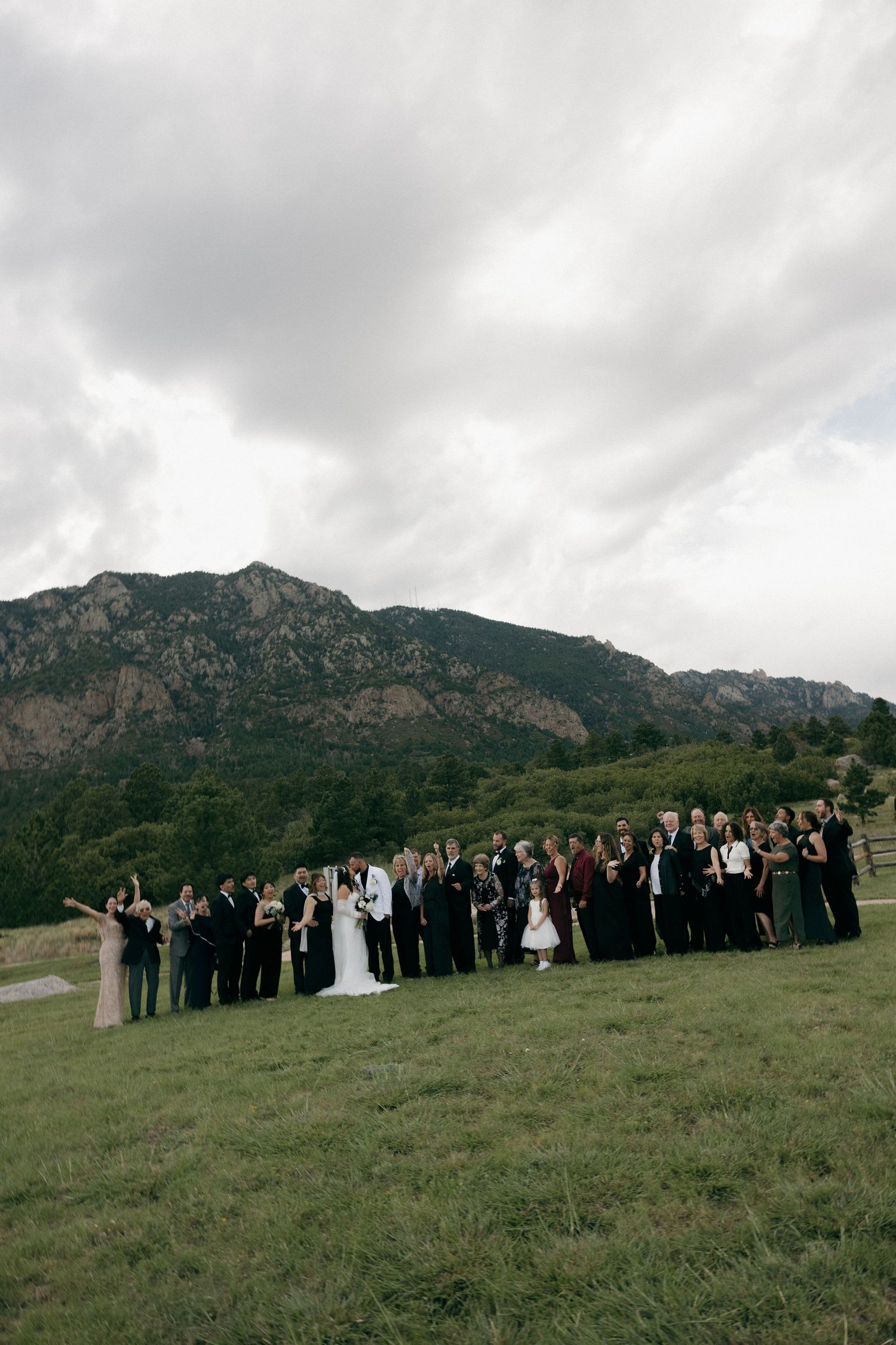 Large wedding party and guests pose together on a grassy hillside with mountain views.