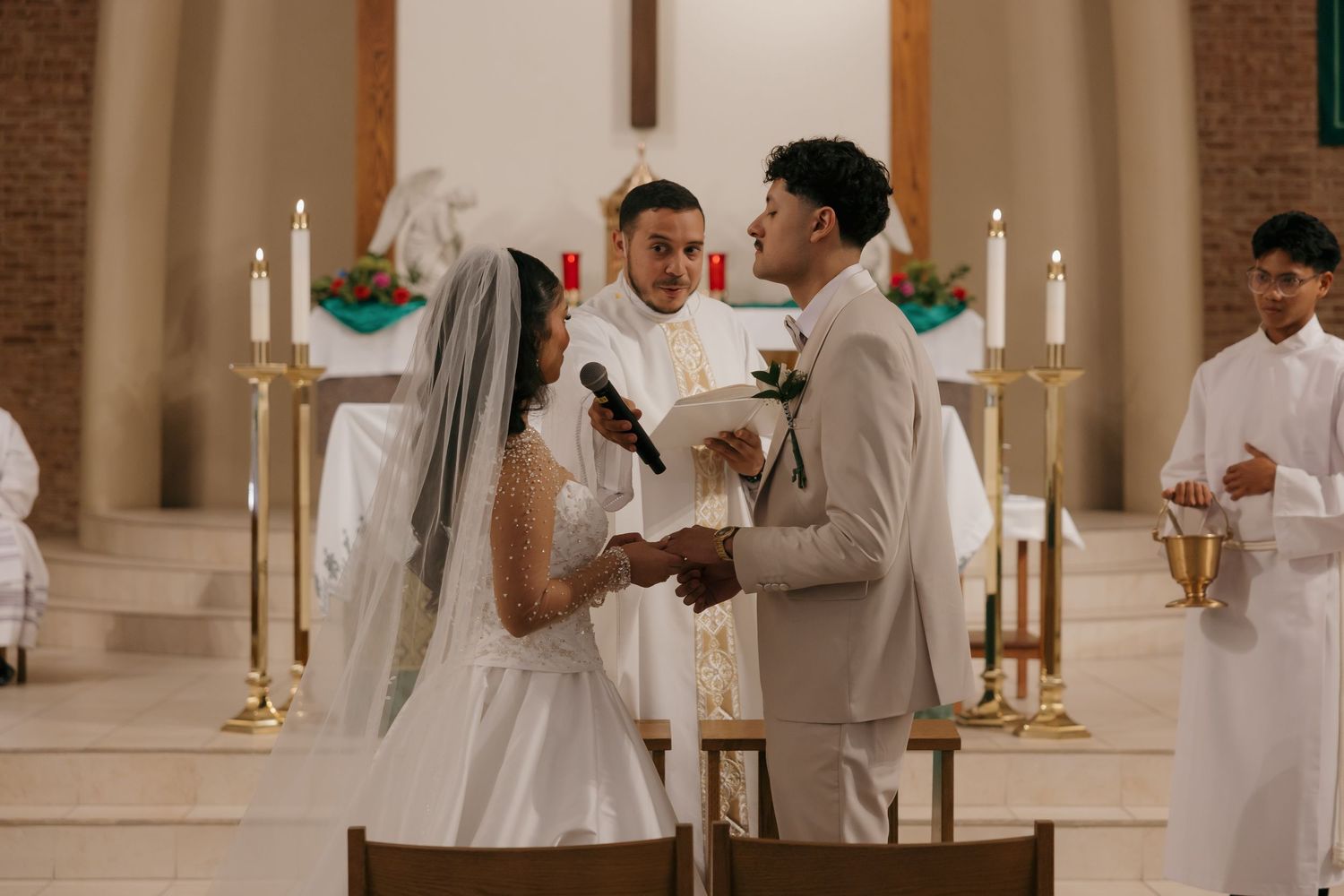 Bride and groom exchange vows at church altar with priest officiating during elegant Catholic wedding ceremony.