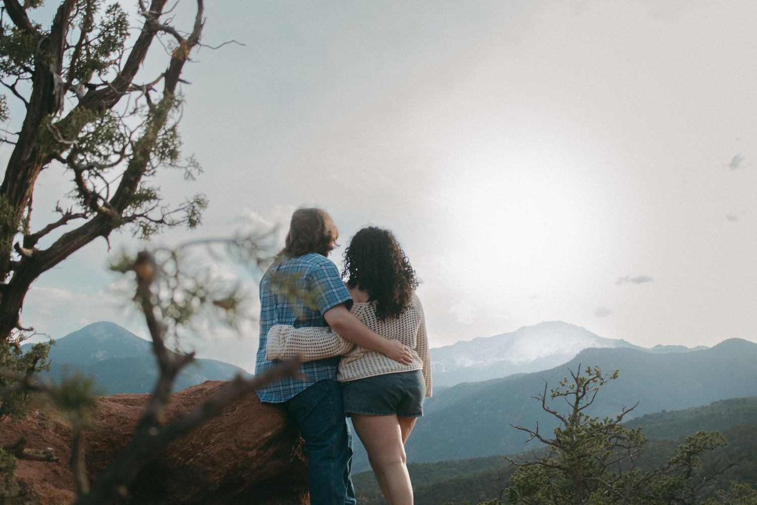 Couple stands together on rocky overlook with blue mountains in the distance.