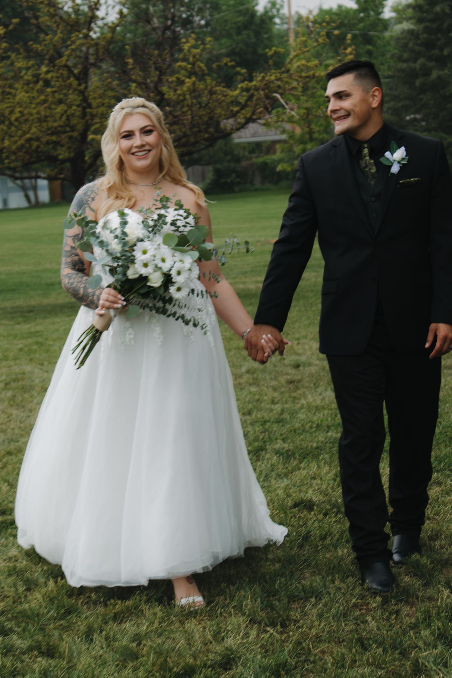 Wedding party members pose together on grass with white floral bouquets and formal attire.