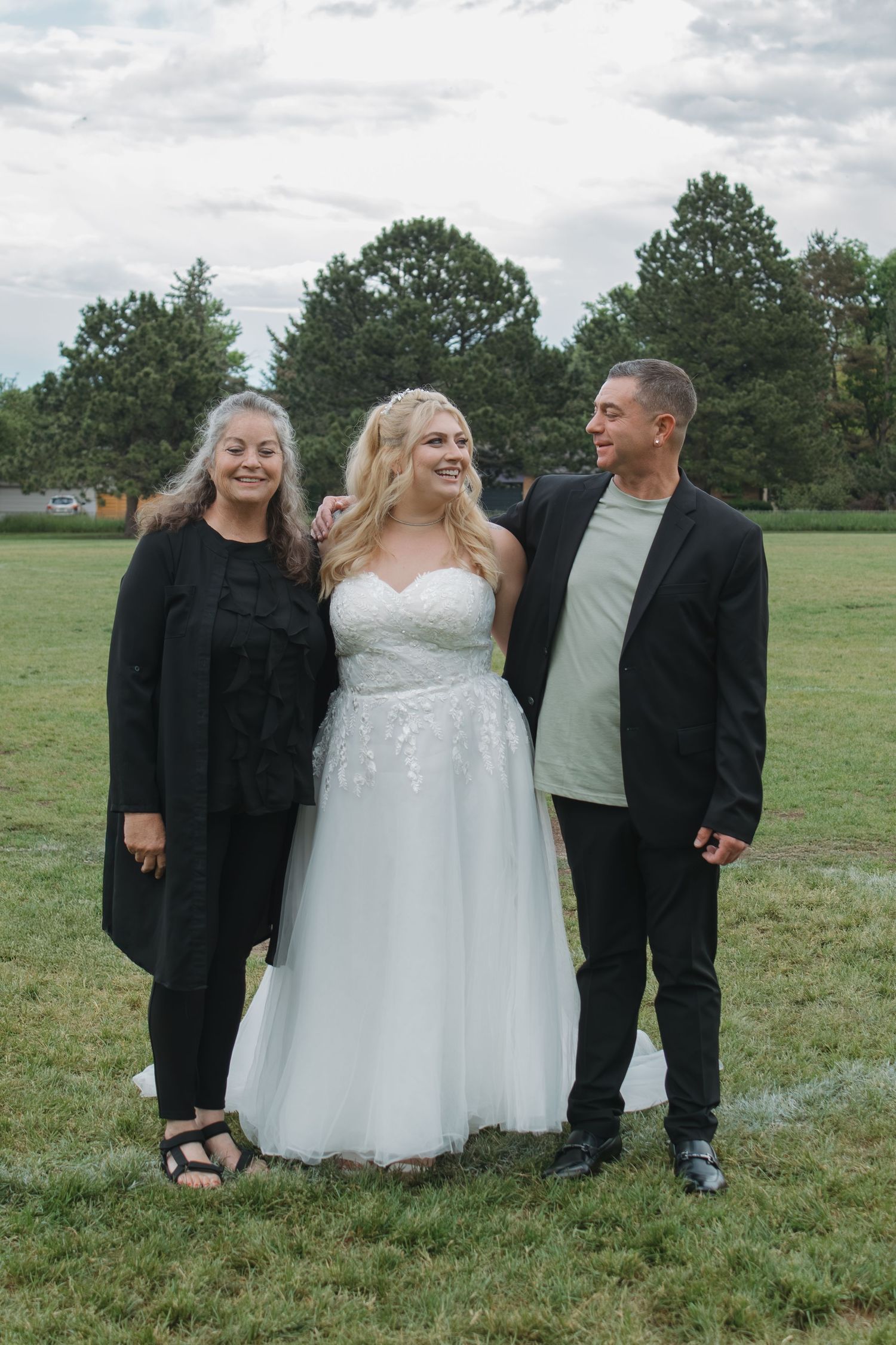 Wedding group poses casually together on grassy field under cloudy sky.