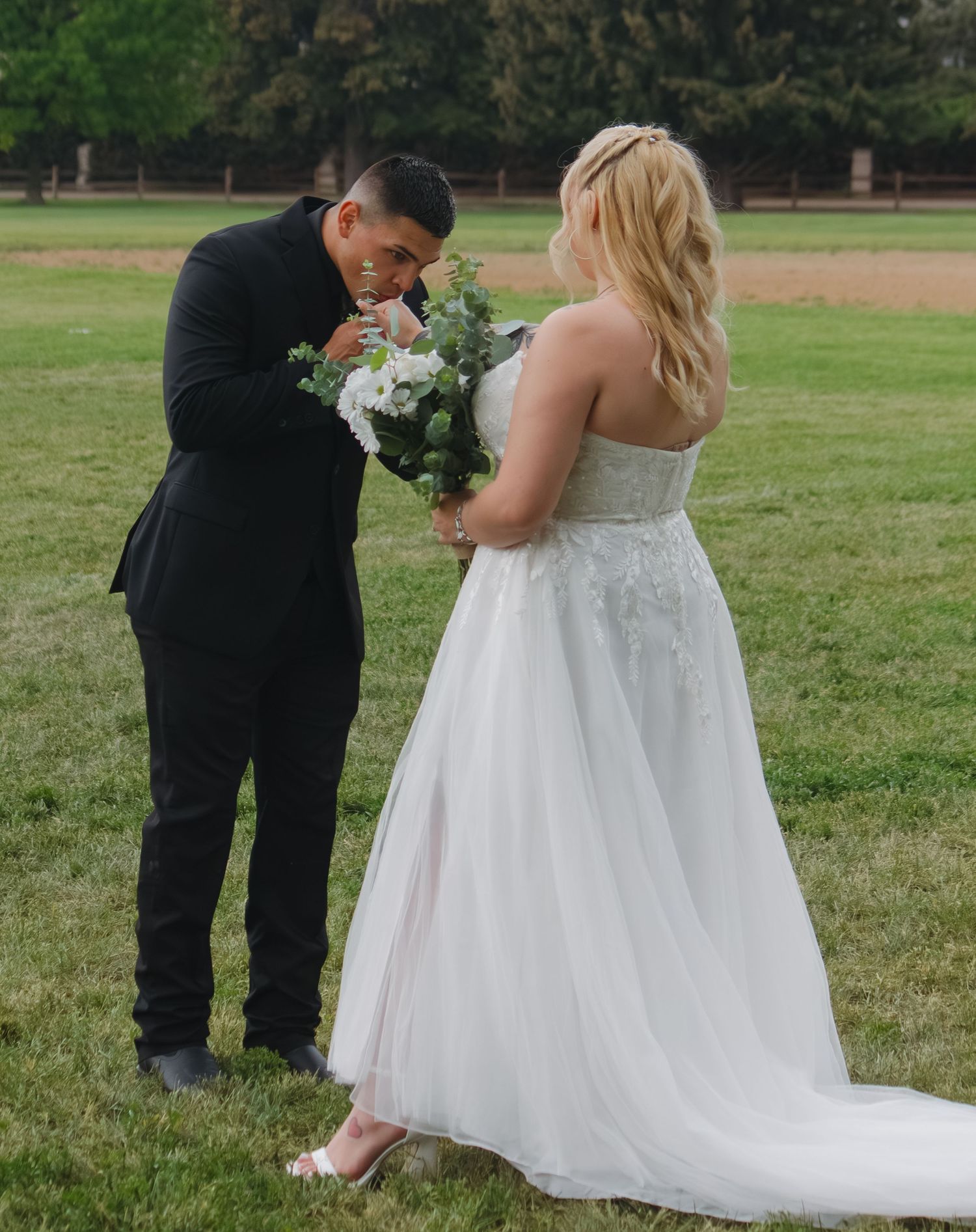 Groom smelling bride's white flower bouquet in an outdoor field setting.