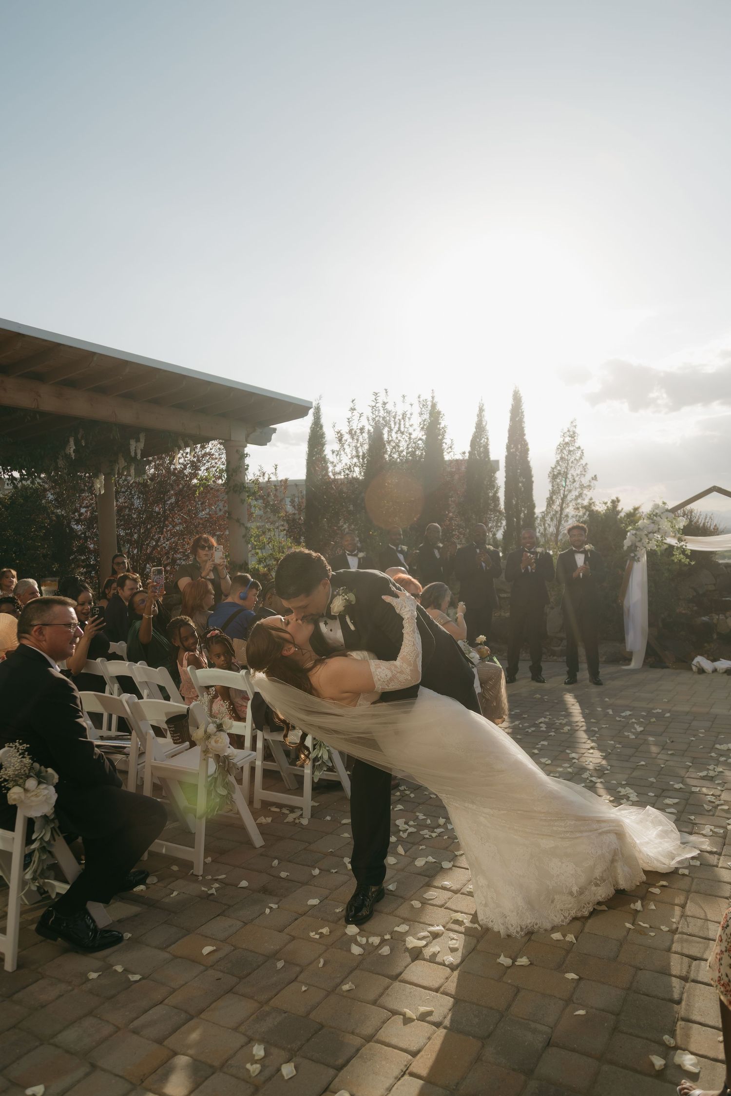 Groom dipping bride for a kiss at outdoor wedding ceremony with guests and sunflare.