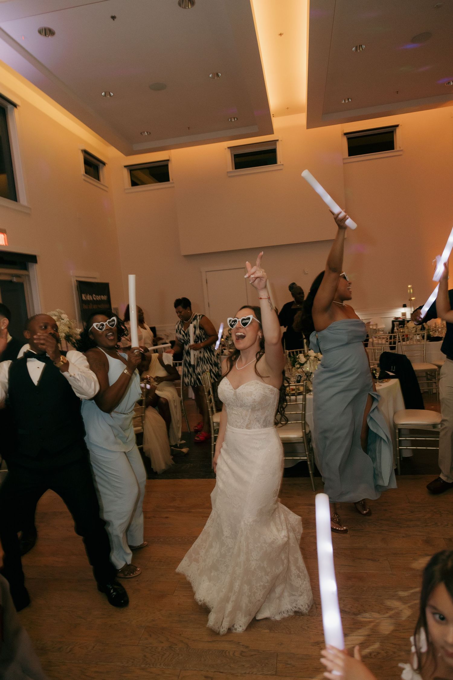 Joyful bride dancing with glow sticks at wedding reception wearing sunglasses with guests celebrating.