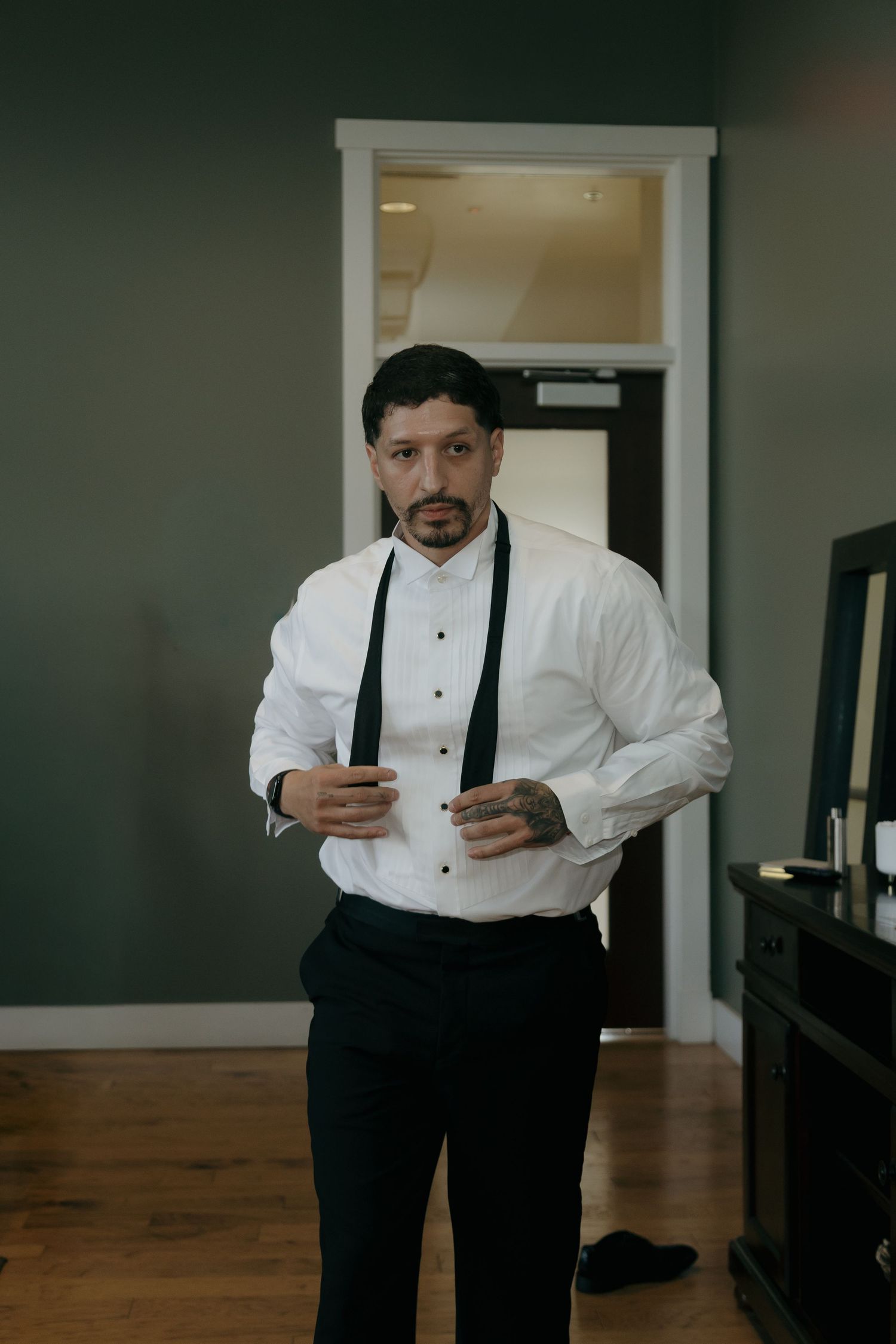 A groom in a white dress shirt and black suspenders adjusts his cufflinks while getting ready for his wedding.