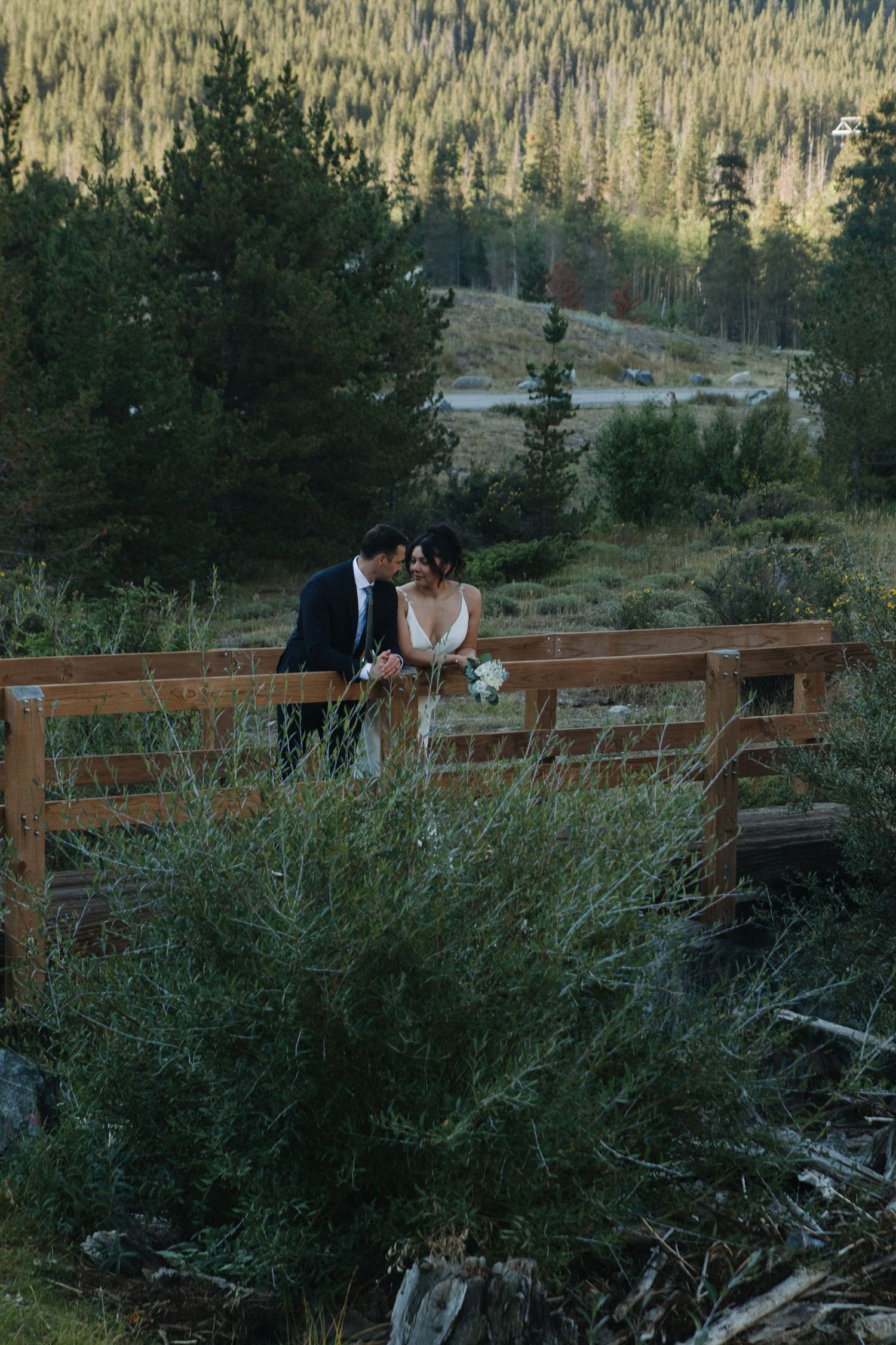 A couple sits together on a wooden bridge surrounded by mountain scenery.