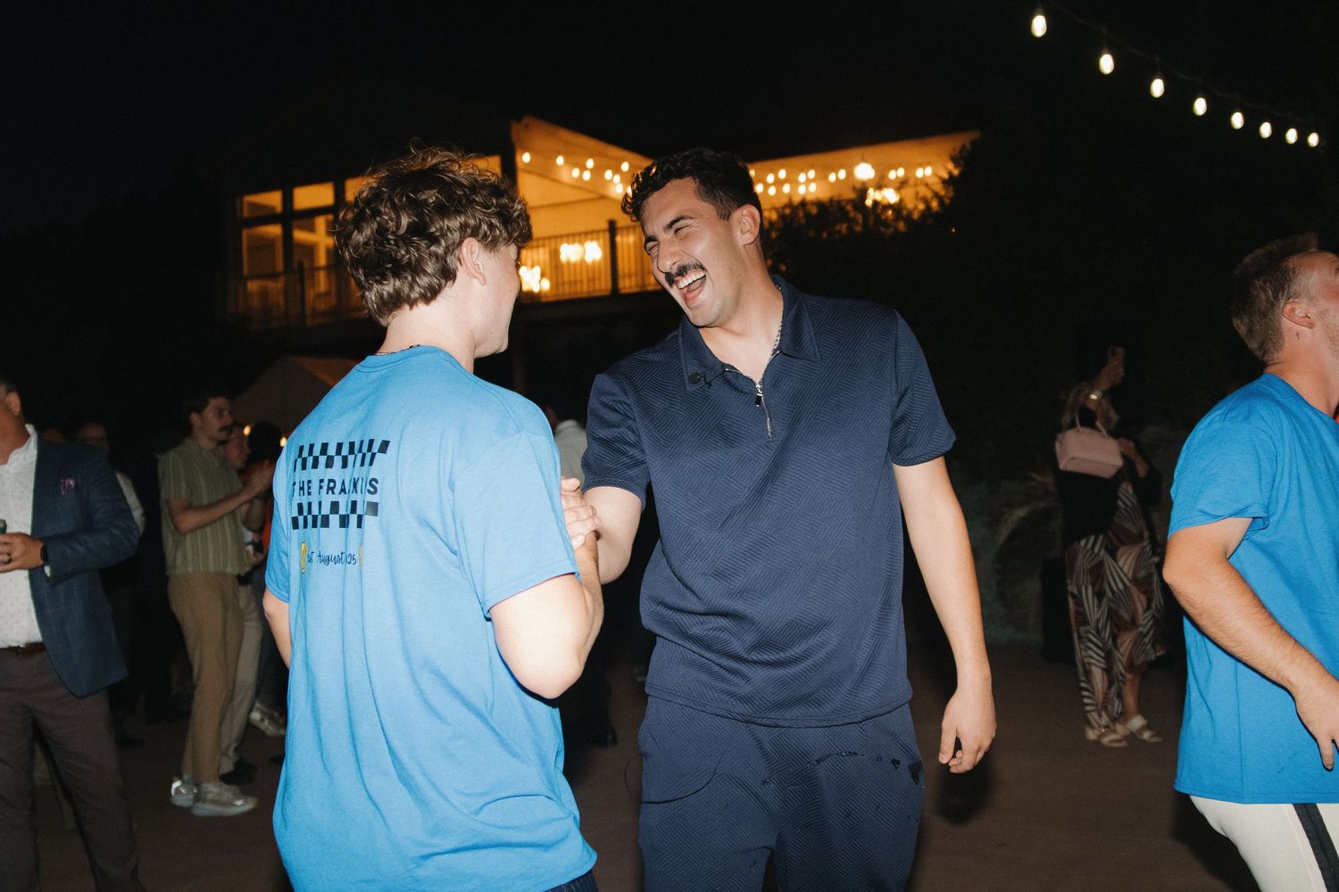 Two people in casual clothing share a laugh while standing at an outdoor nighttime gathering with string lights.