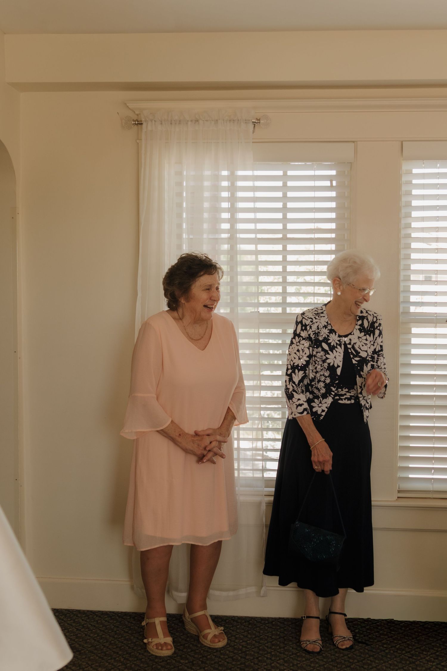 Two elderly women in contrasting dresses sitting together against a wall with shuttered windows.