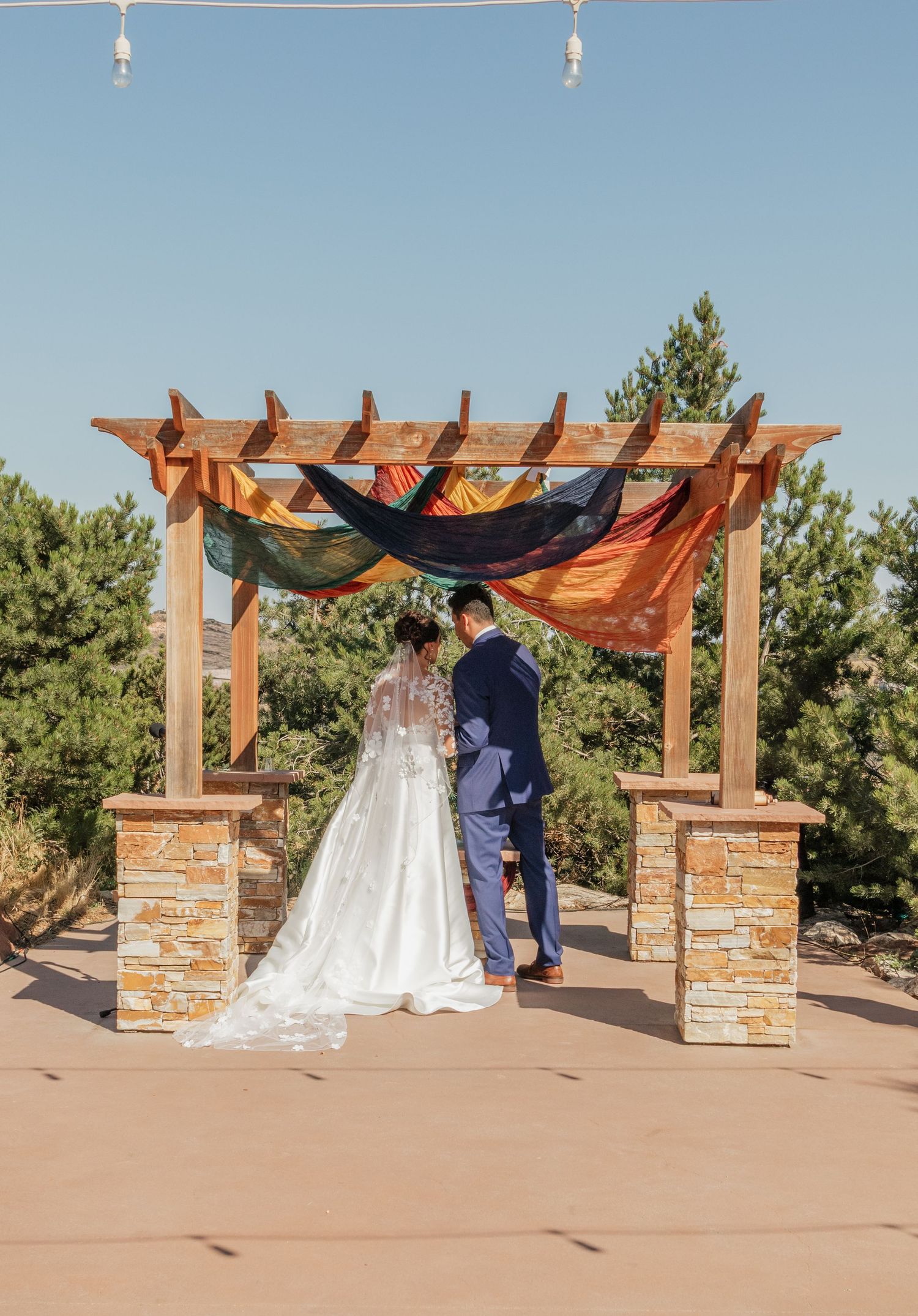 A wedding couple shares a romantic moment under a rustic wooden pergola with stone pillars in an outdoor setting.