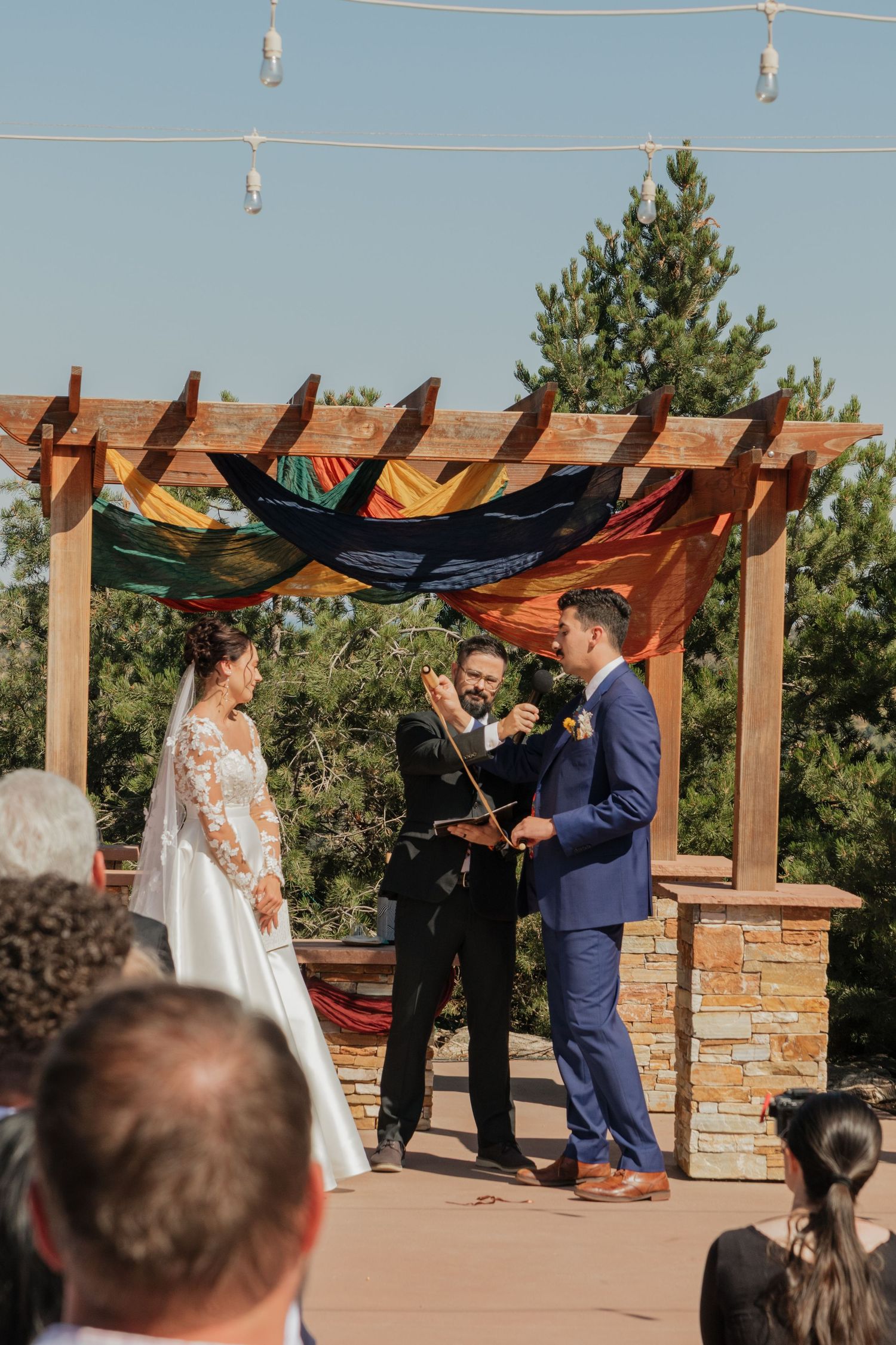 An outdoor wedding ceremony with guests seated beneath a wooden pergola structure.