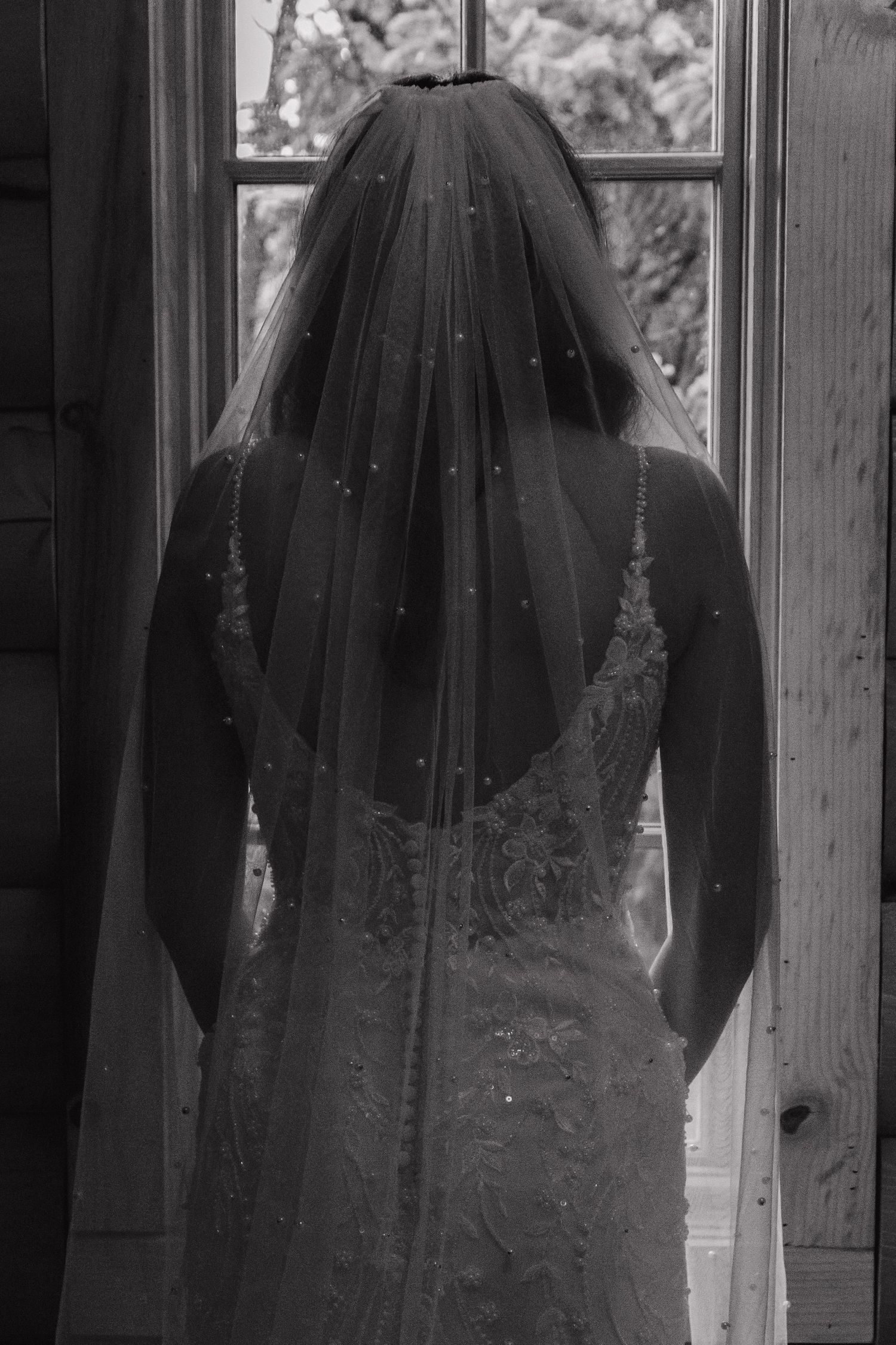 Black and white photo of a bride in a lace wedding dress with a long veil standing by a window.