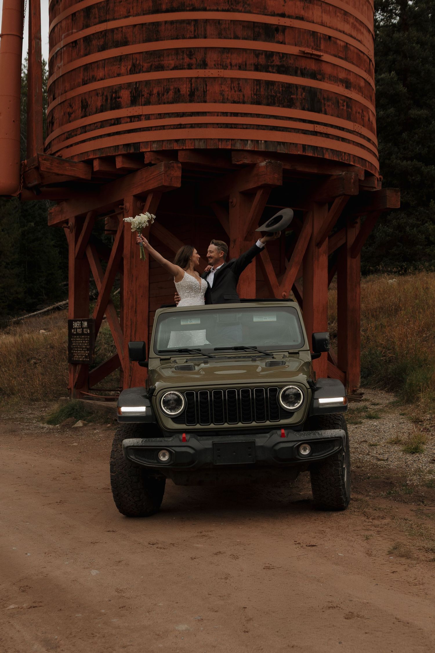 A couple poses in a black Jeep beneath large wooden structures in a rustic mountain setting.