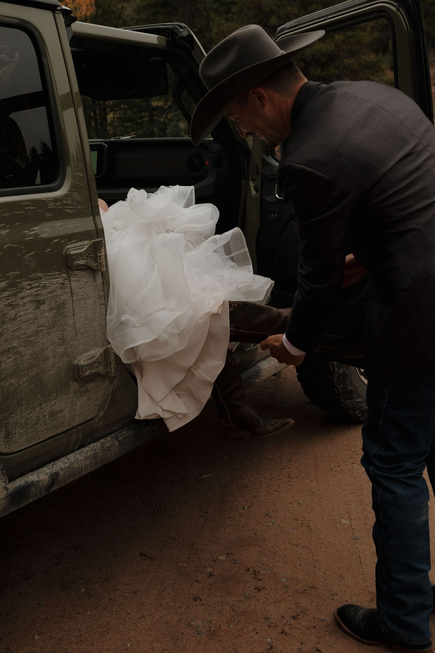 A cowboy helps with a wedding dress near a vehicle door.