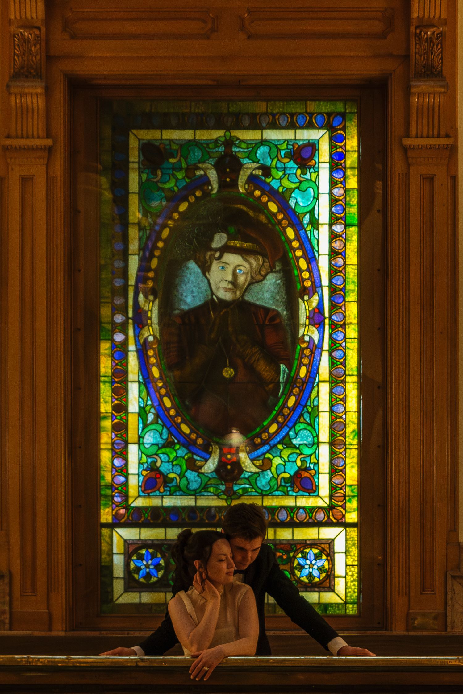 A couple shares an intimate moment in front of ornate stained glass windows with rich golden lighting.
