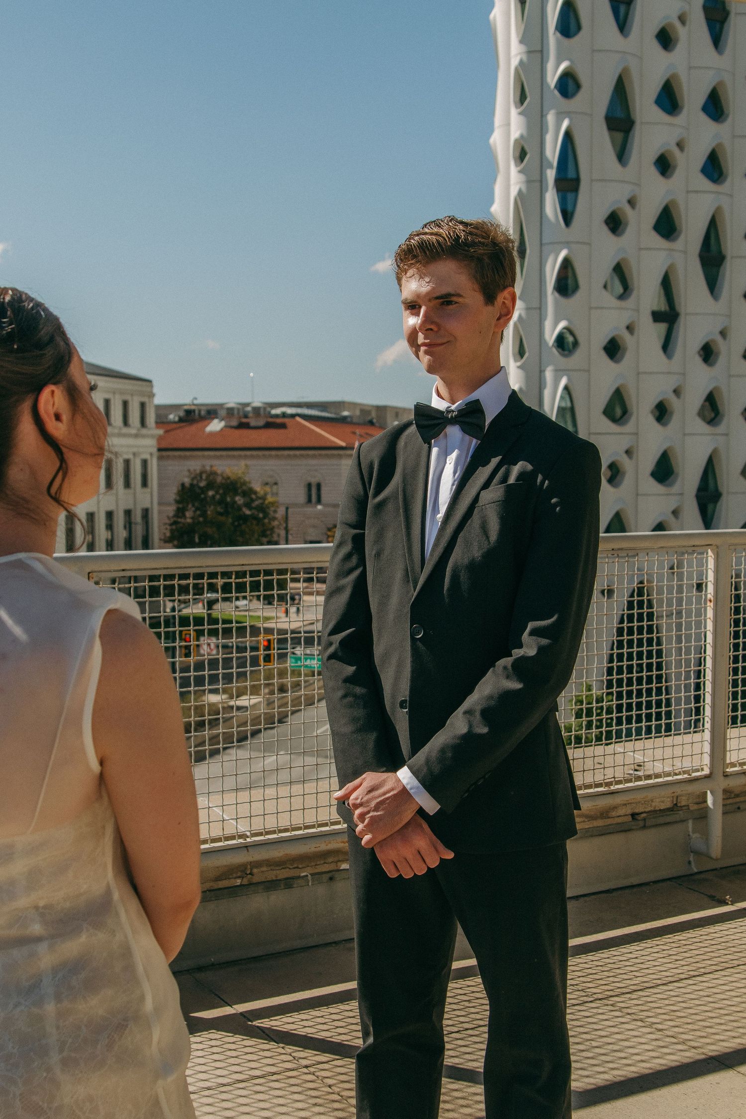 A couple in formal attire stands on a balcony with a modern architectural backdrop.