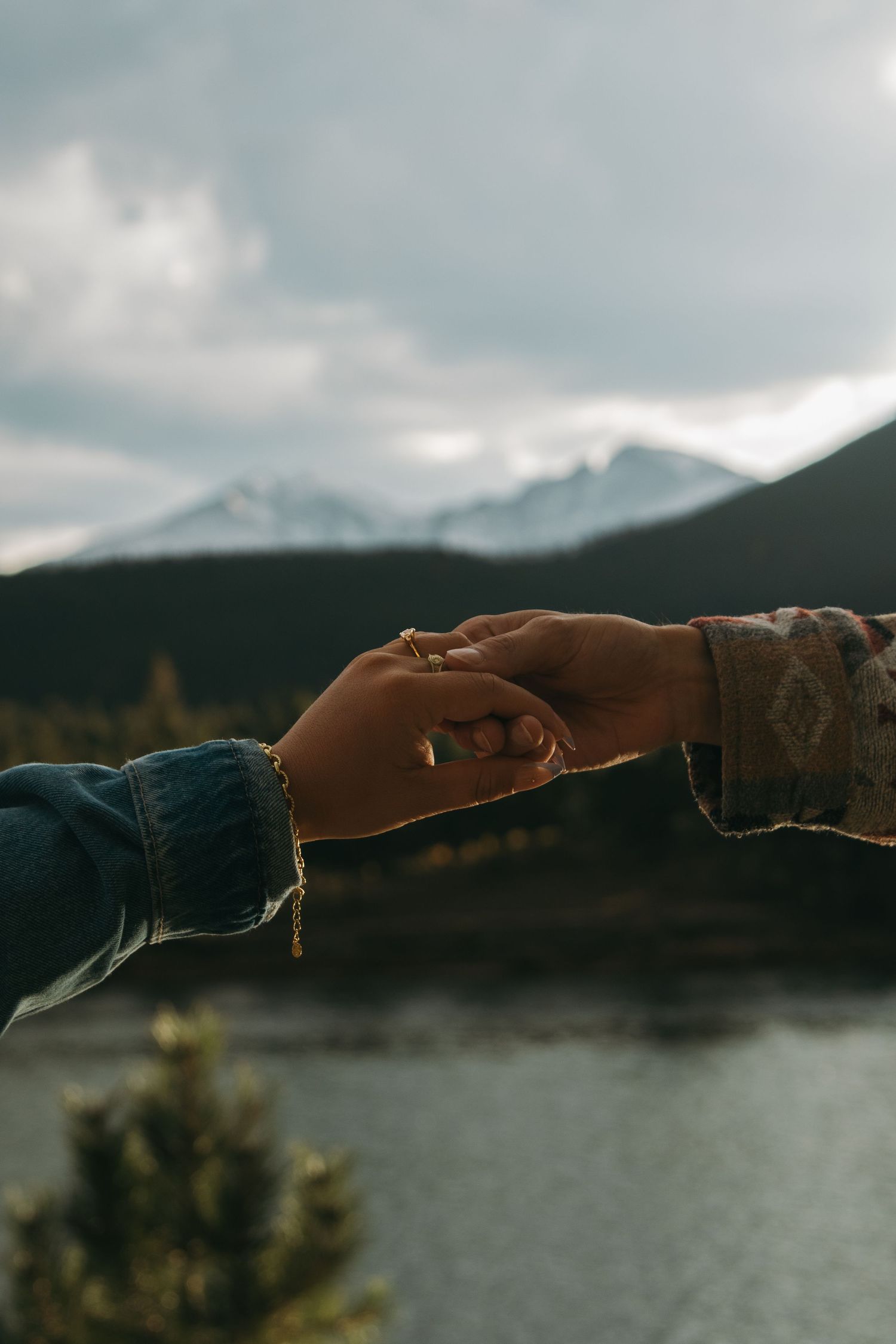 Two hands reaching out to hold each other against a scenic mountain landscape with a lake in the background.