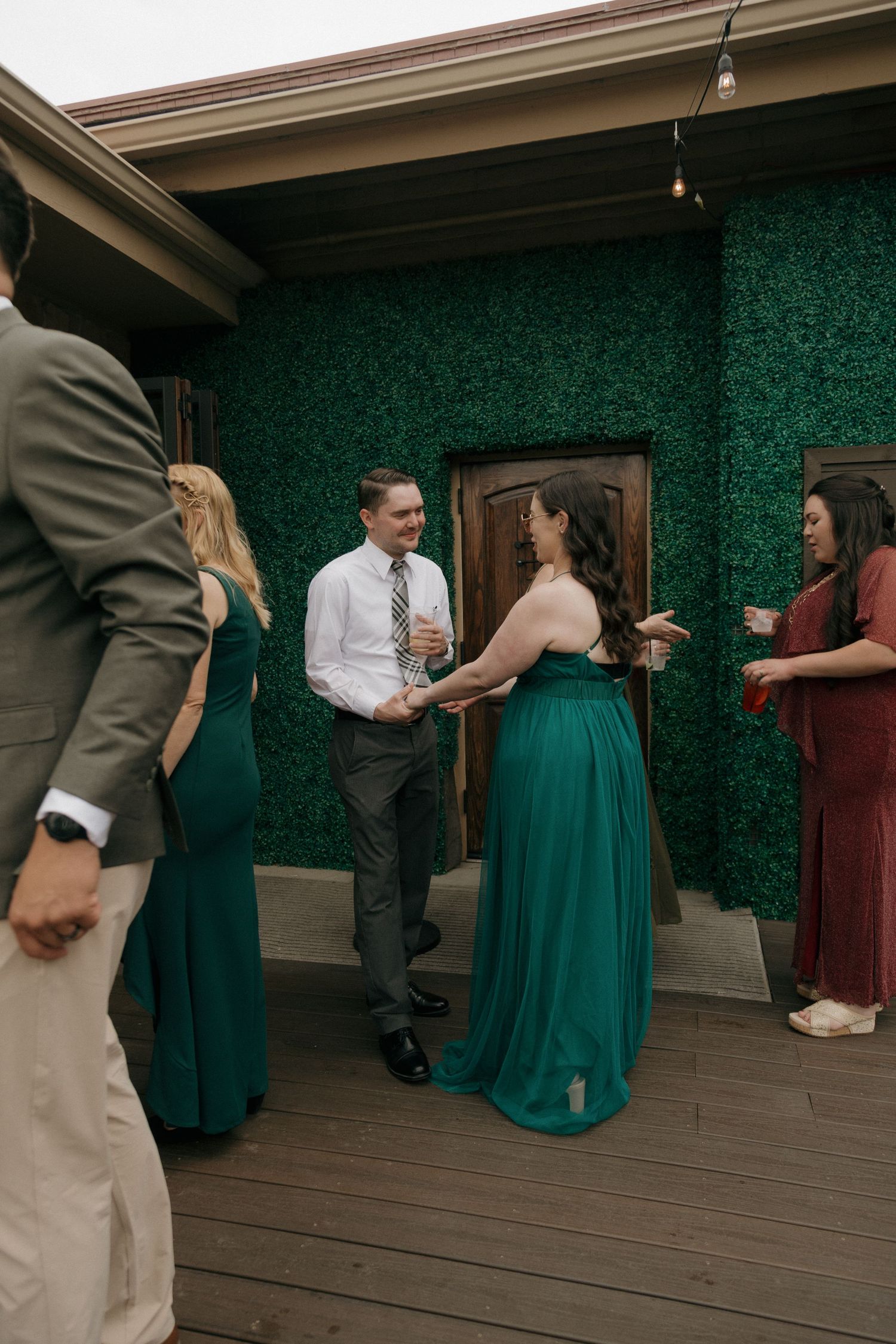 Wedding guests in emerald green dresses mingle on a deck with a green hedge wall backdrop.