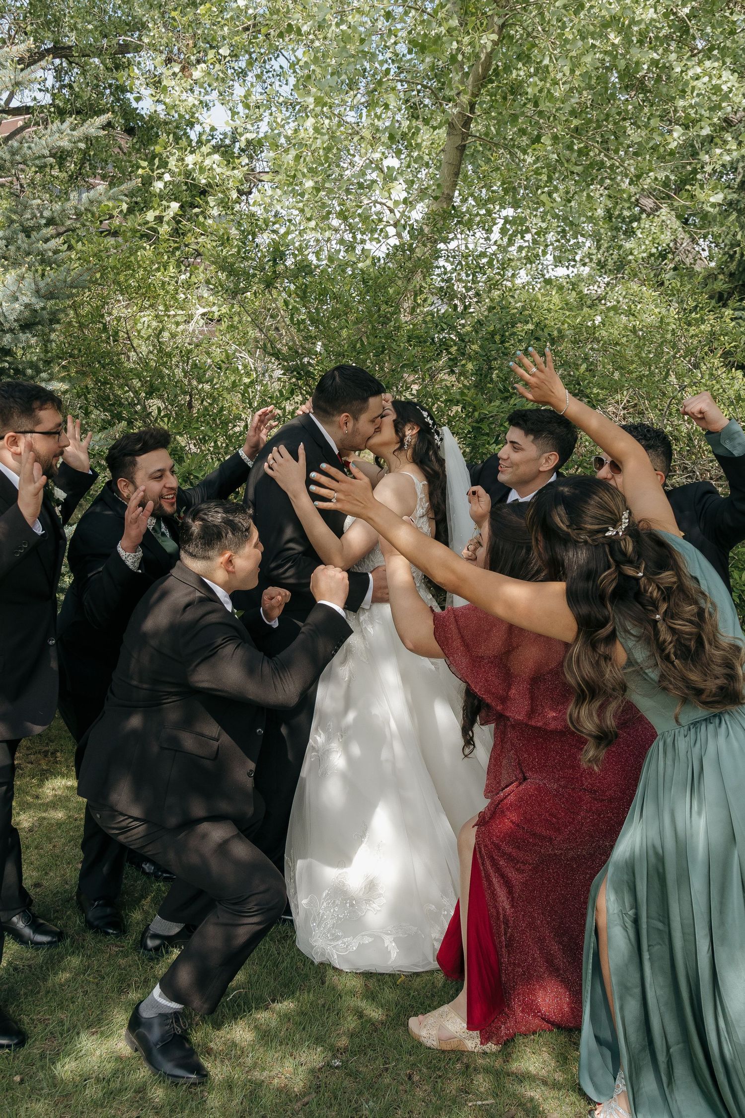 Wedding party celebrates bride and groom with cheers and raised hands under sunlit green trees outdoors.