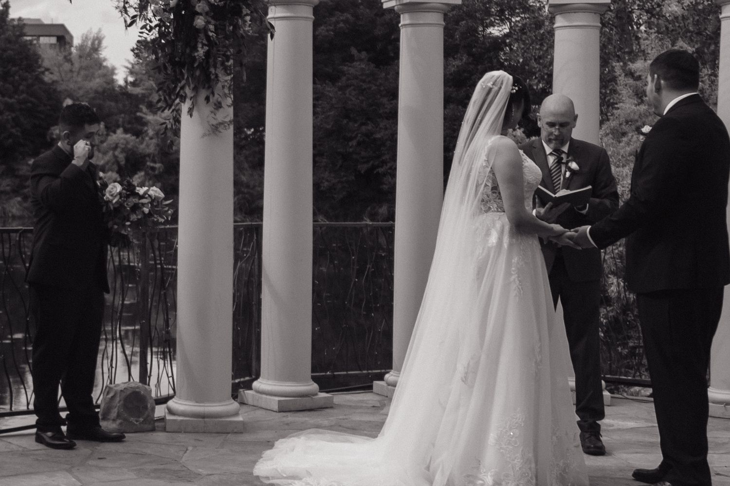Bride and groom exchange vows at outdoor wedding ceremony with elegant white columns in black and white.