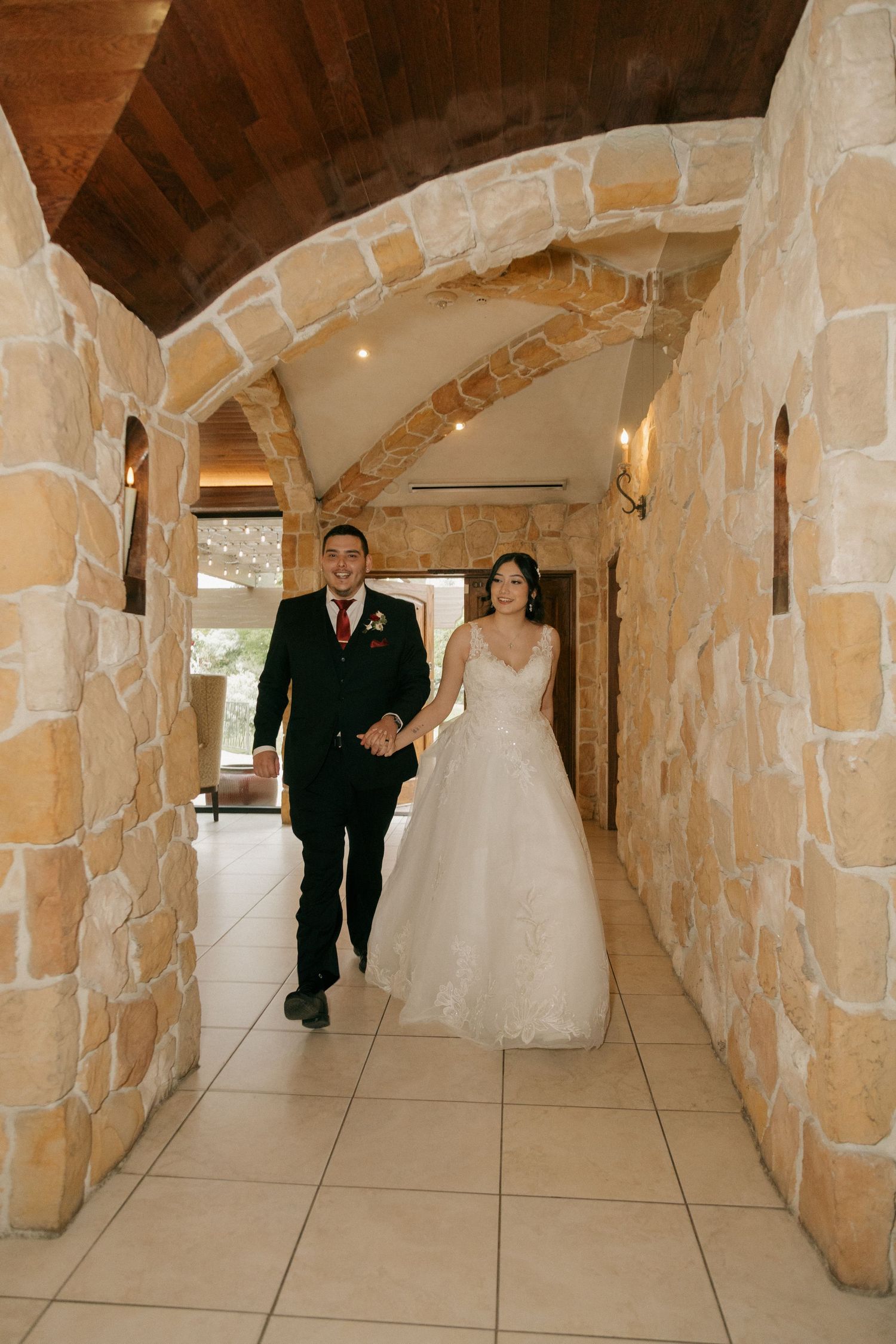 Bride and groom walk hand-in-hand through a rustic stone archway corridor at their wedding venue.