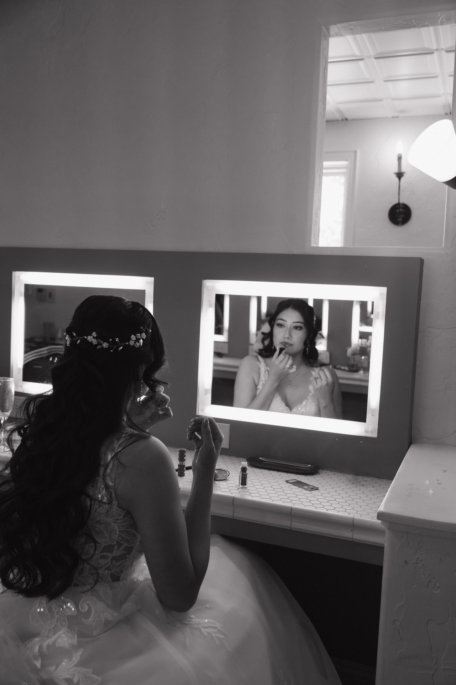 Bride applying makeup at a vanity mirror with Hollywood lights during wedding preparations.