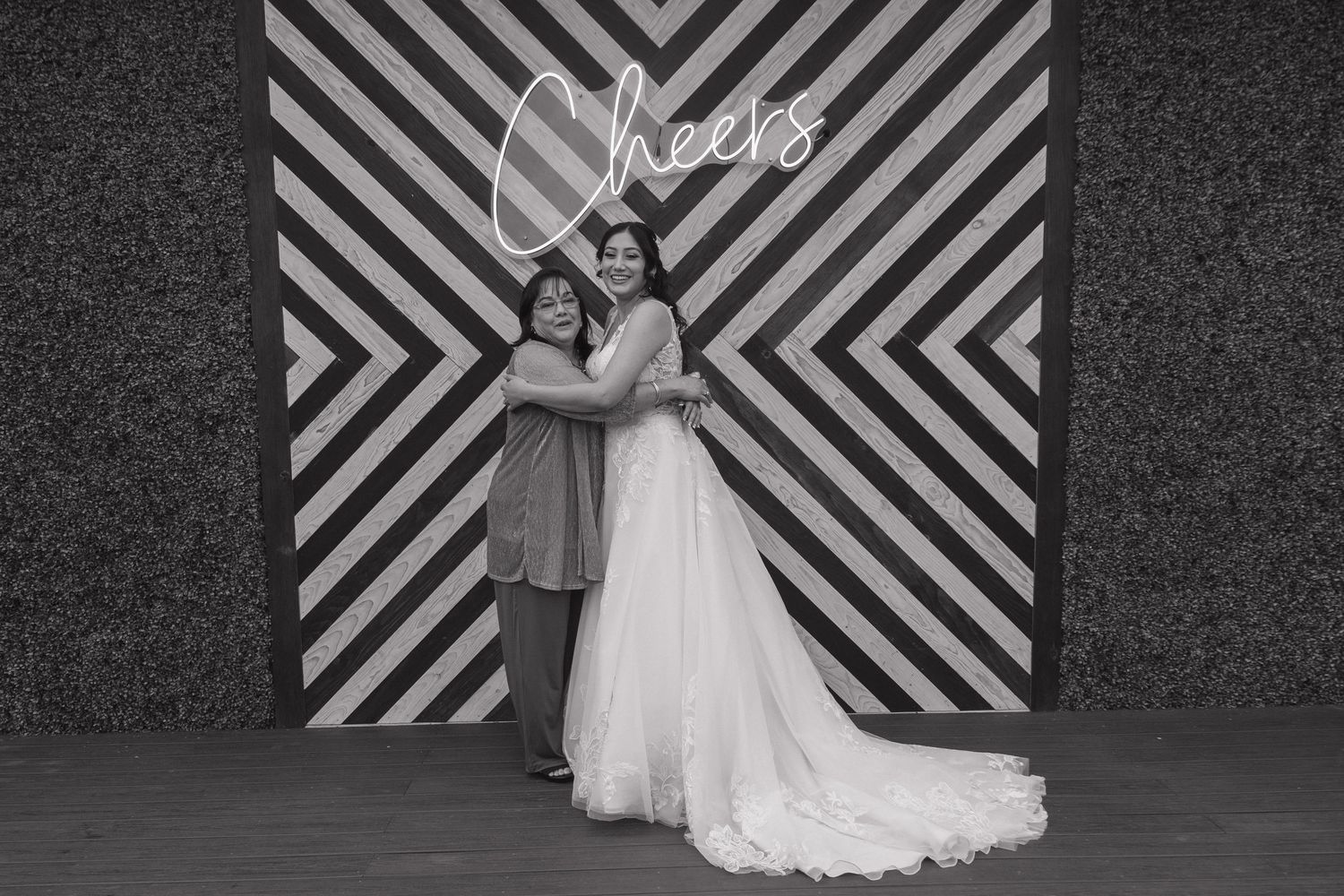 Black and white photo of bride in white gown hugging guest in front of Cheers backdrop.