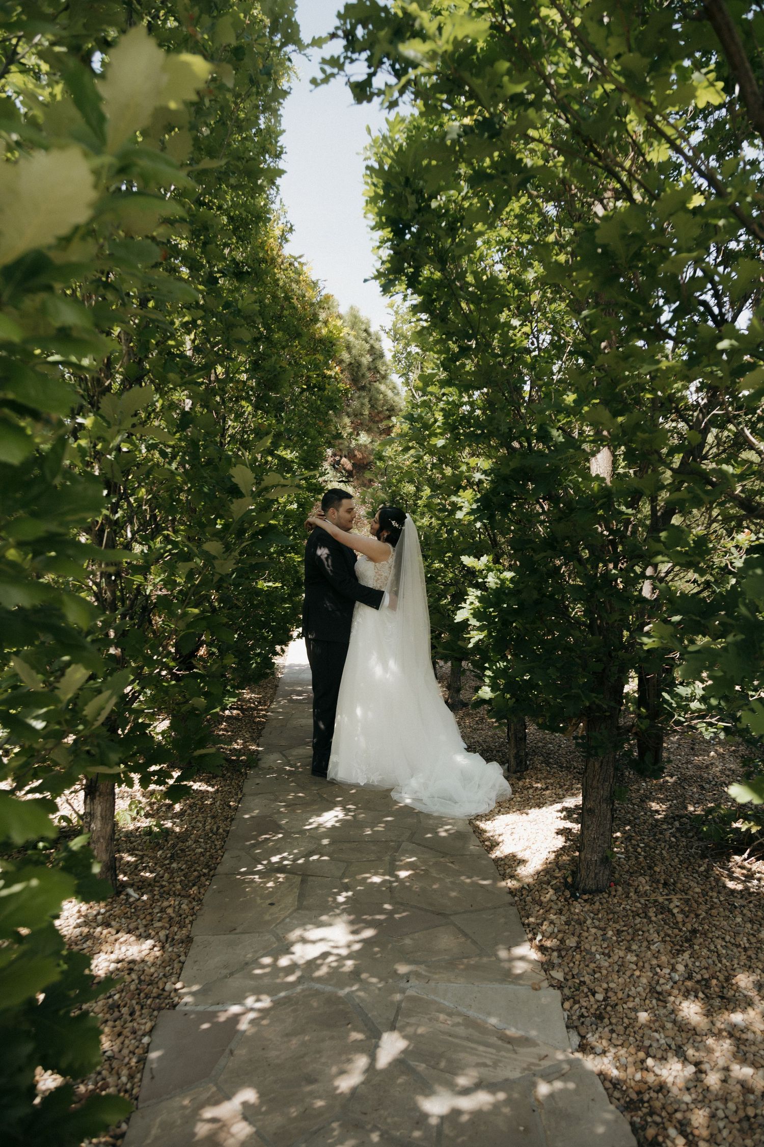 Bride and groom sharing romantic moment on stone pathway surrounded by lush green trees.