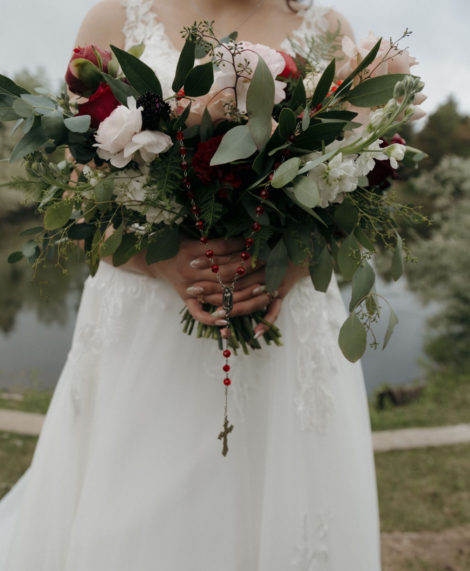 Bride holds a lush bouquet of white and red flowers with a red rosary draped over her hands.