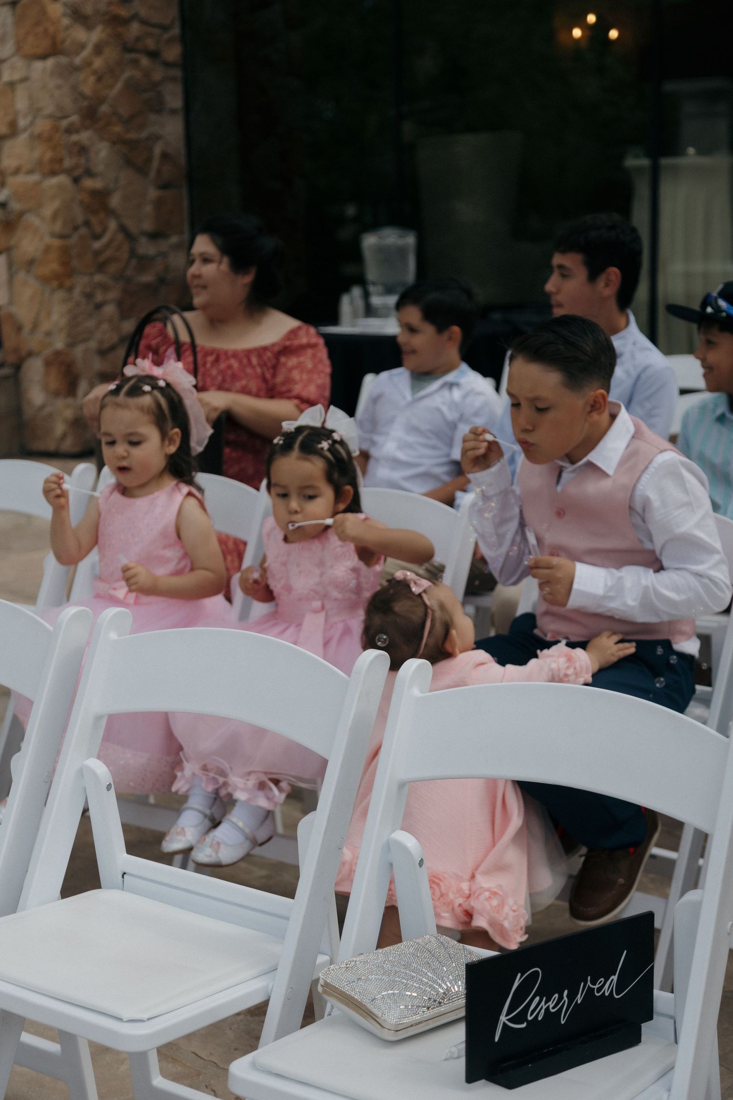 Young children in pink dresses sit in white chairs at outdoor wedding ceremony near Reserved sign.