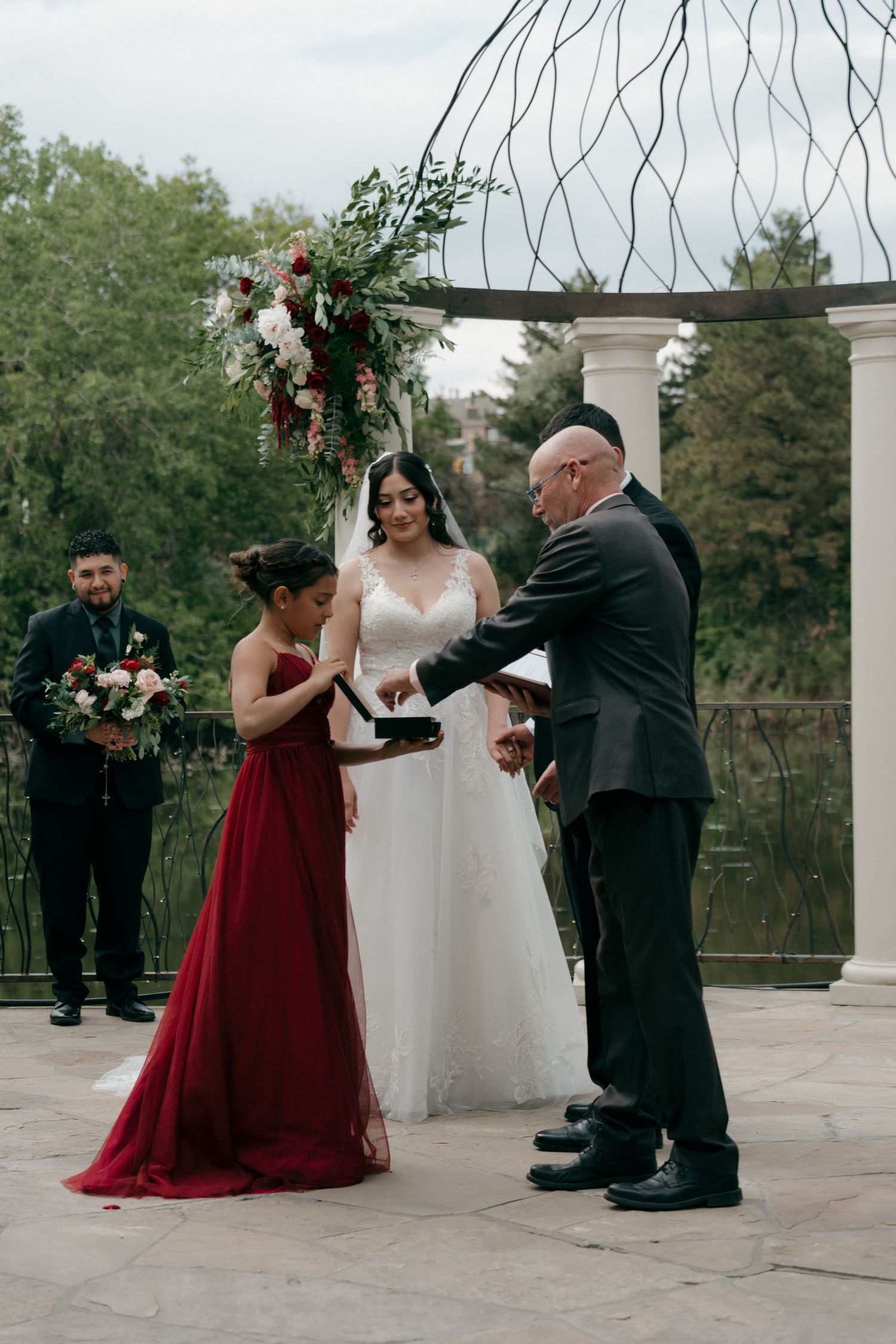 Bride receives wedding ring from officiant as bridesmaid in red gown assists at outdoor ceremony.