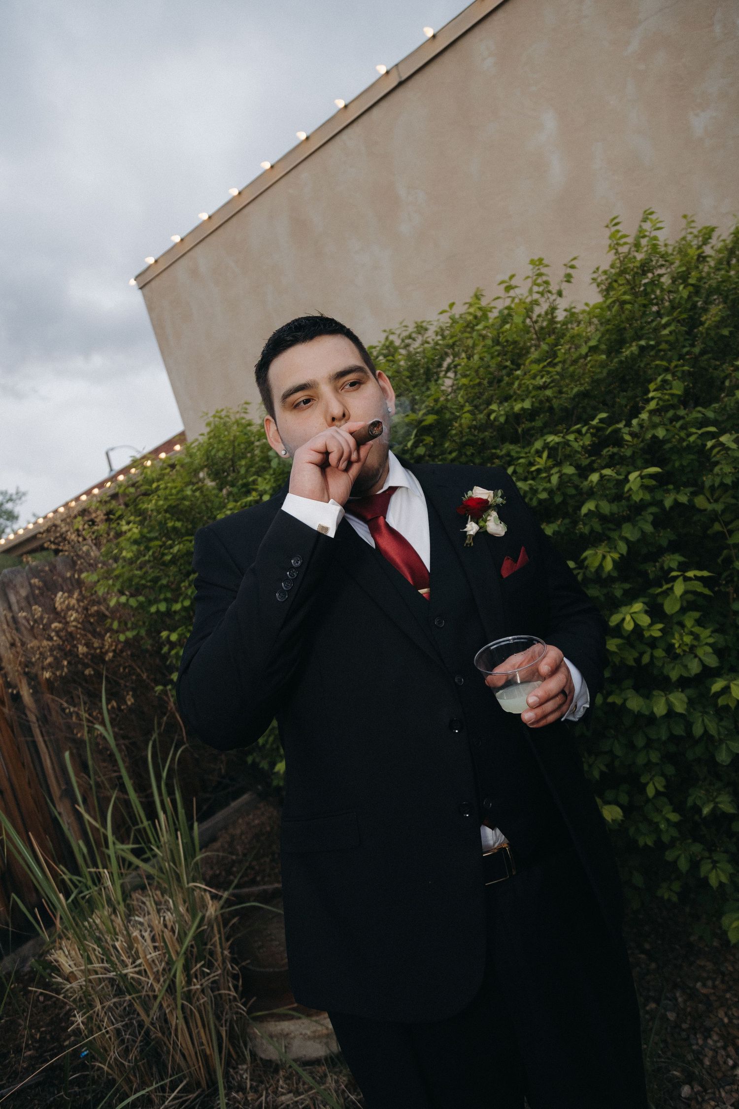 Groom in a black suit smokes a cigar and holds a drink outdoors after the wedding.