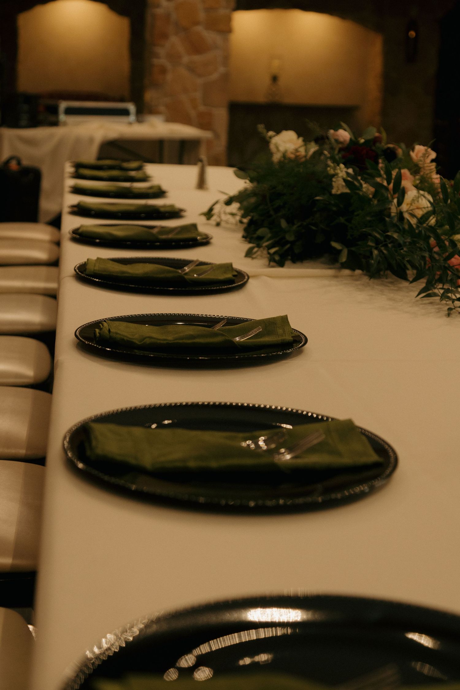 Elegant wedding reception table setting with dark green charger plates, olive napkins, and floral centerpieces on a white tablecloth.