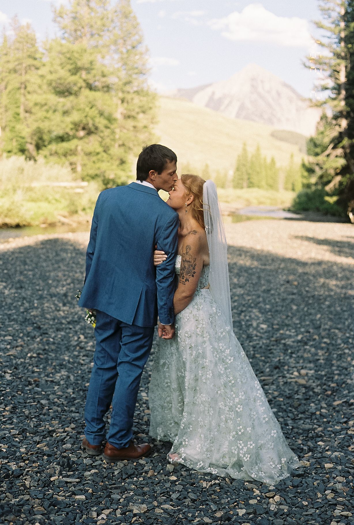 Newlyweds sharing a kiss in lace wedding dress and blue suit with scenic mountain landscape.