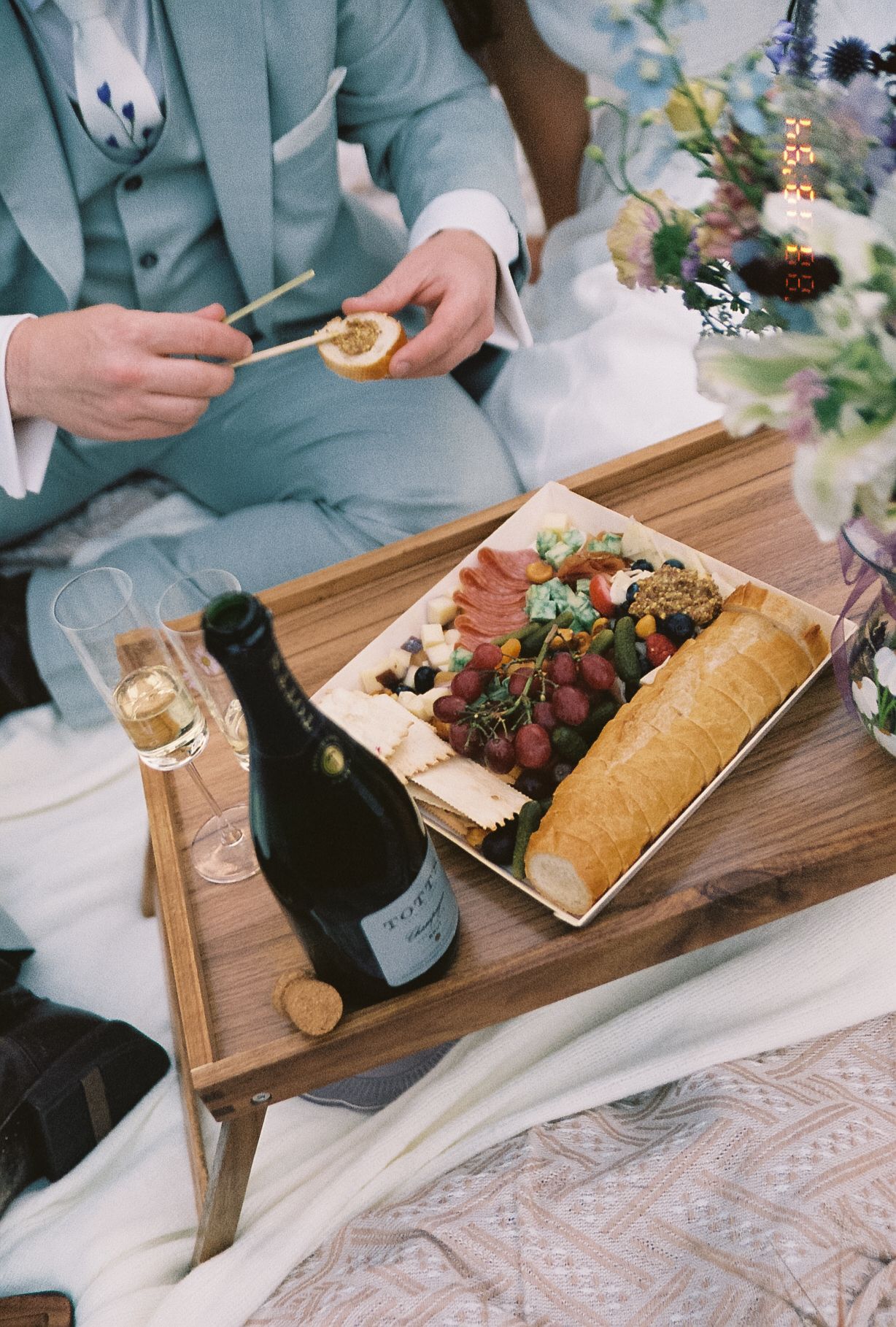 Groom enjoying charcuterie board with baguette grapes and champagne on wooden tray during outdoor picnic elopement.