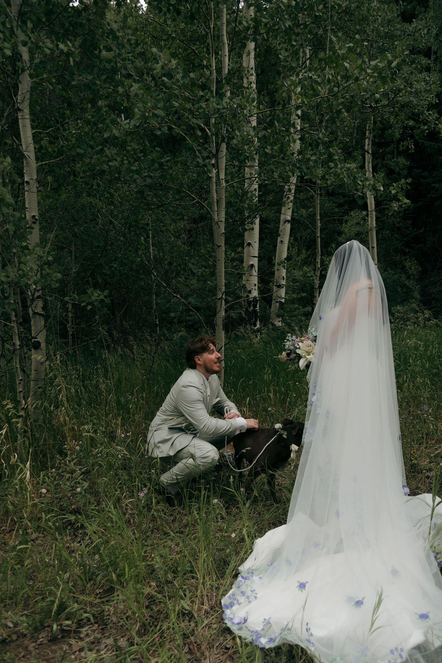 Groom kneeling beside dog in forest during outdoor wedding ceremony with bride in floral veil.