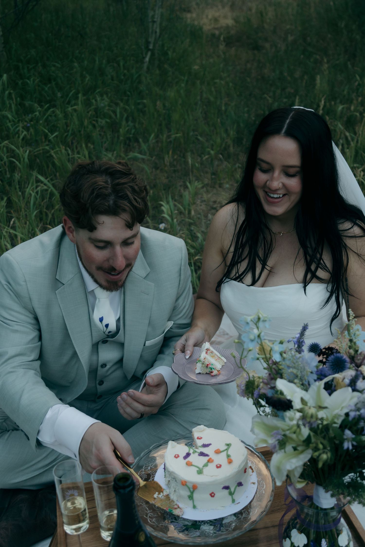 Laughing bride and groom in wedding attire cut a white cake decorated with colorful flowers during an outdoor picnic.
