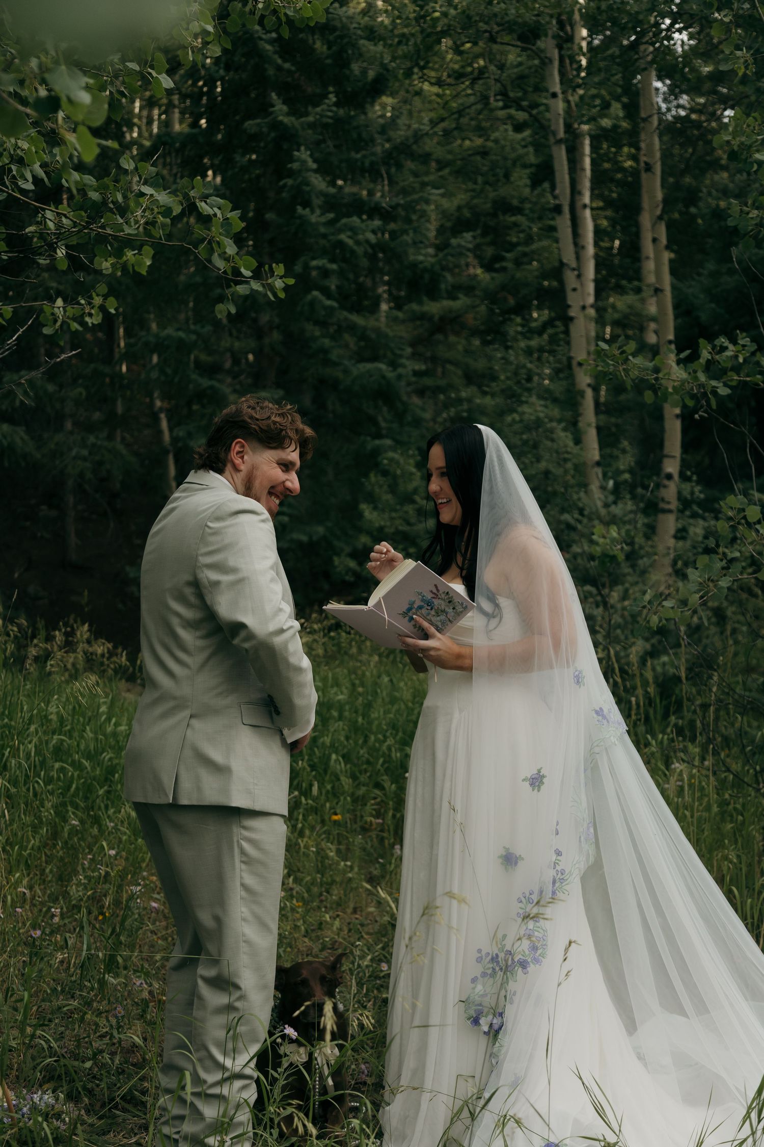 Smiling bride and groom exchange vows from books during a joyful outdoor forest wedding ceremony.