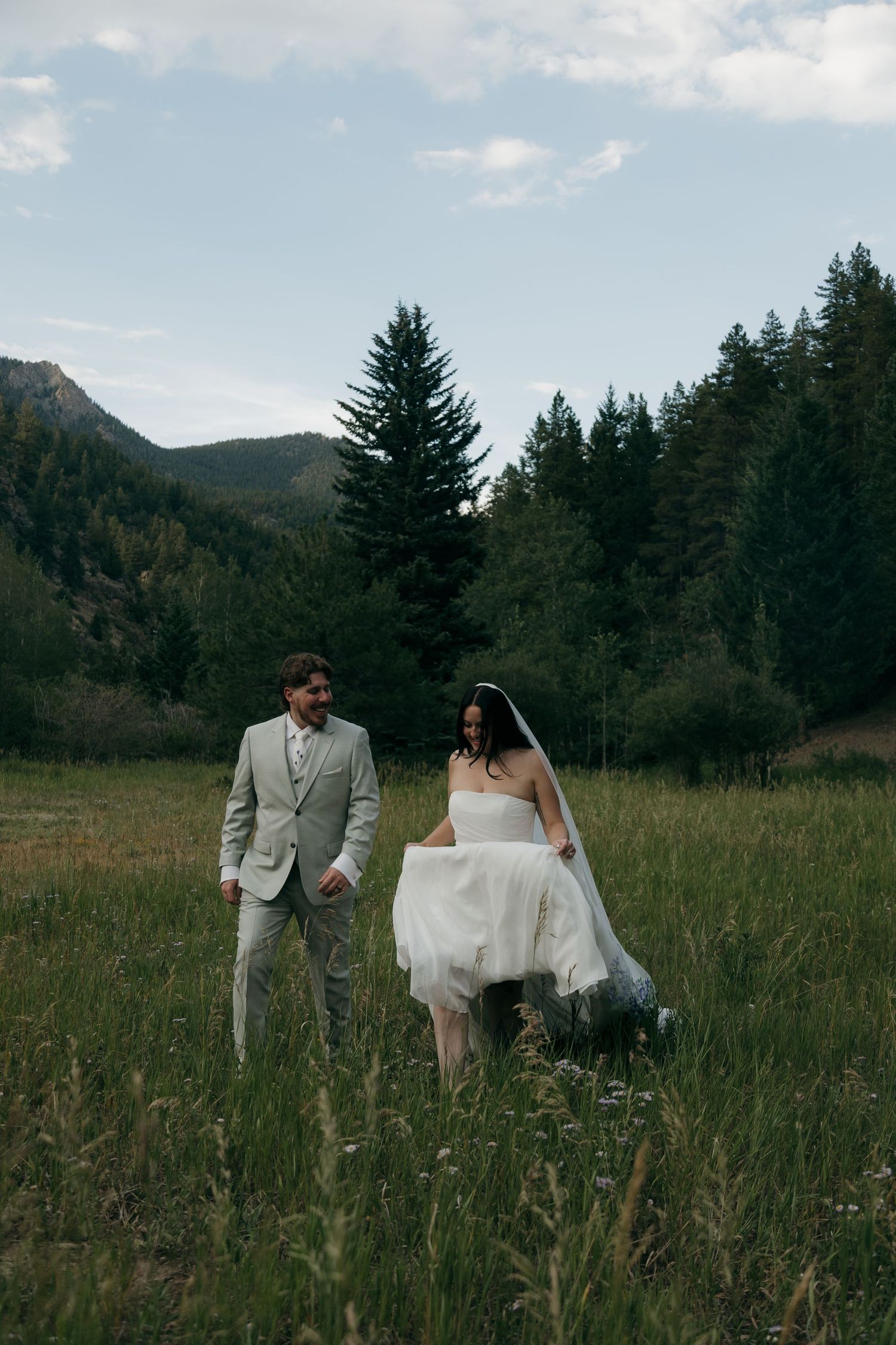 Bride and groom walk through lush green meadow with mountain forest backdrop on wedding day.